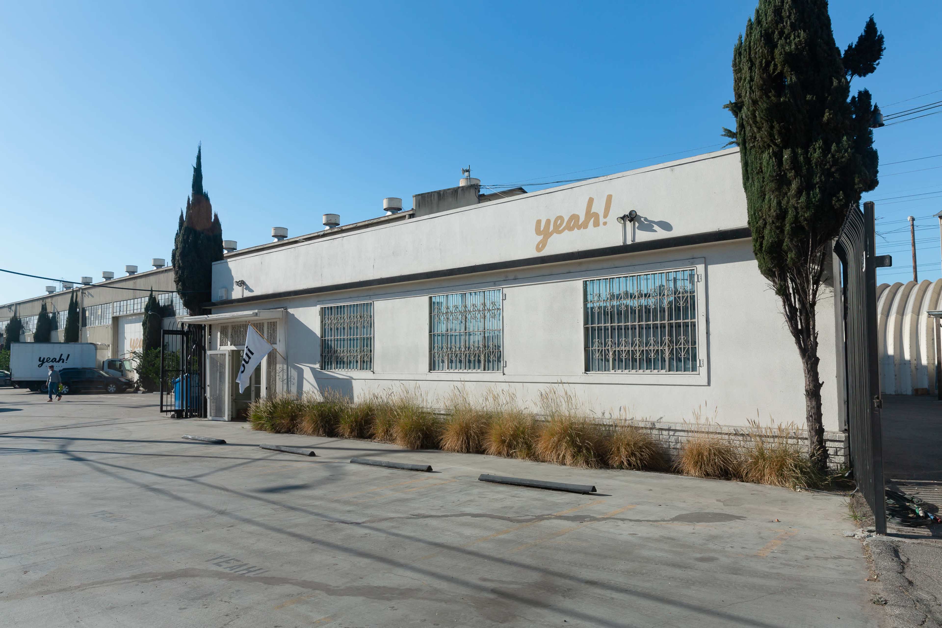 The image shows a white industrial building with the word "yeah!" displayed on its facade, accompanied by neatly trimmed vegetation and a clear blue sky.