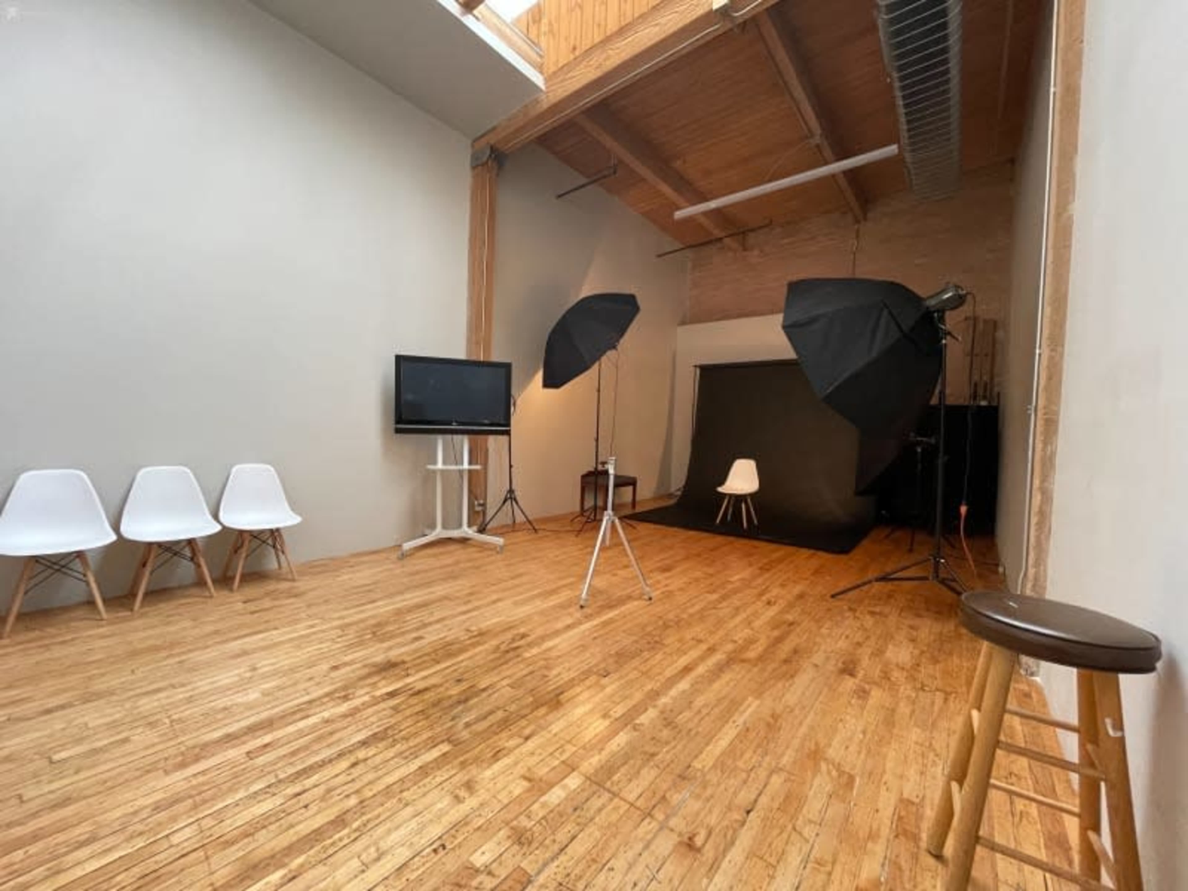 A photography studio with a wooden floor, two large softbox lights, a backdrop, a white chair, and a television on a stand.