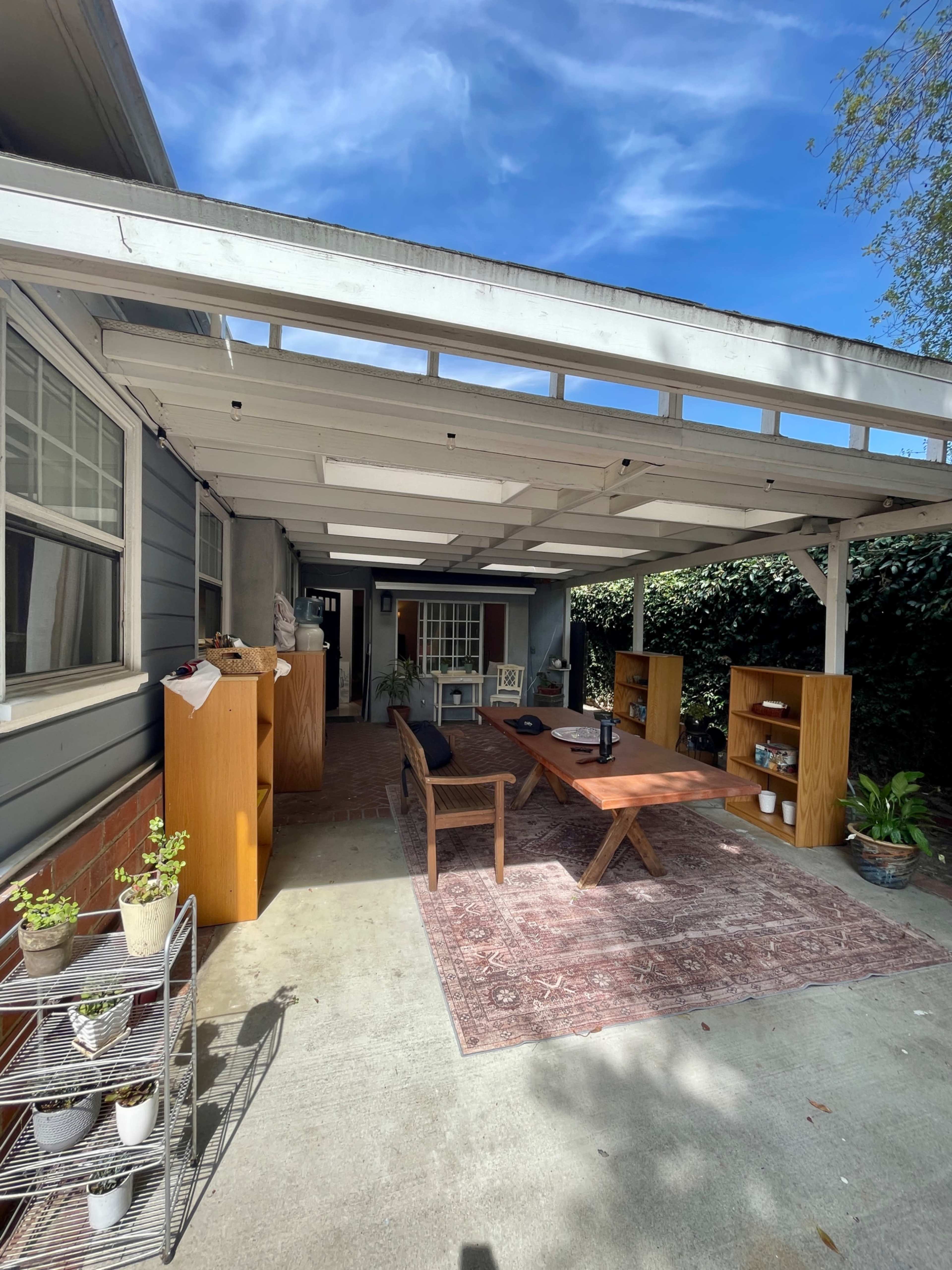 The image shows a covered outdoor patio area with a wooden table, chairs, shelves, and potted plants.