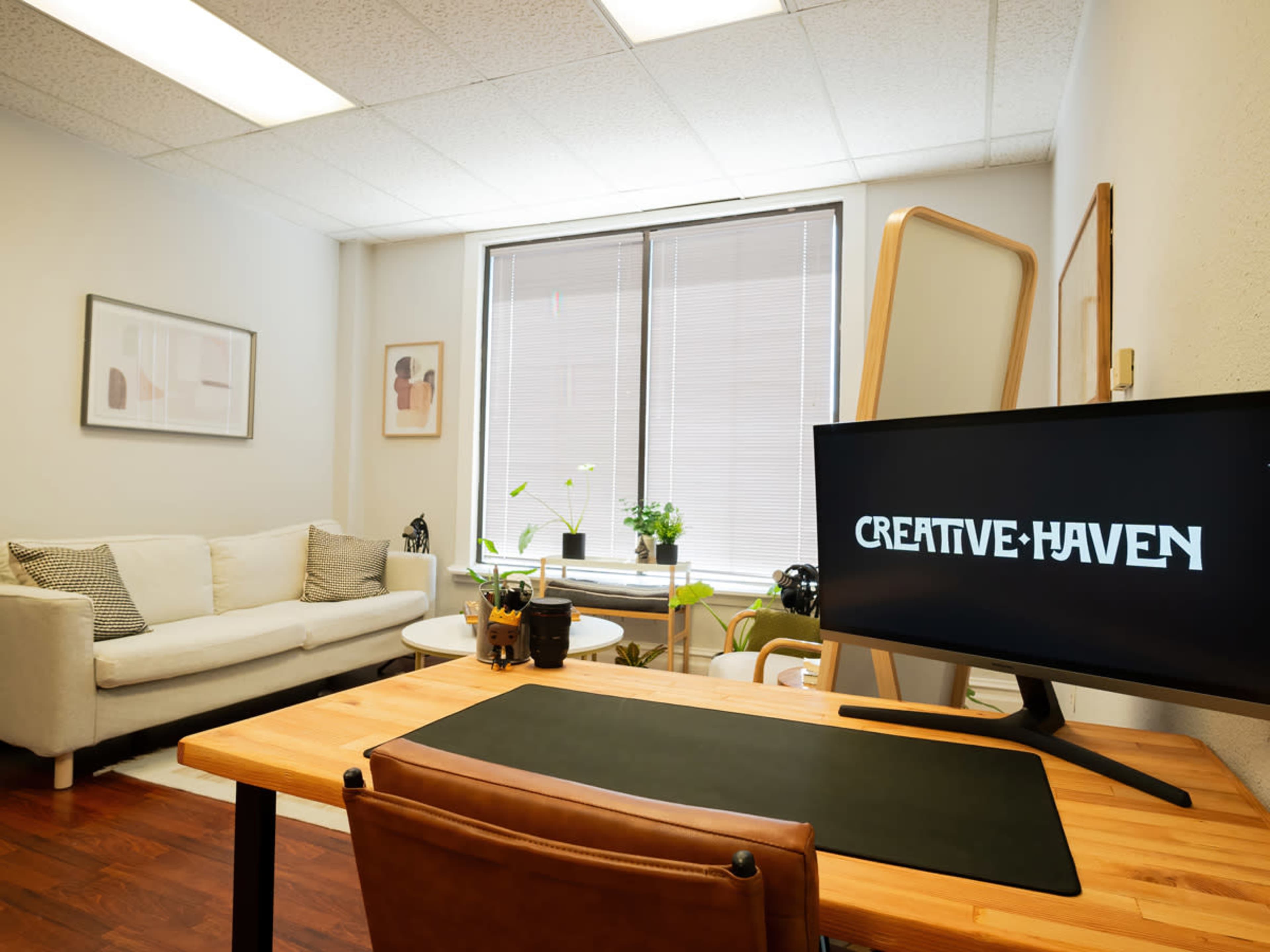 A modern office space with a desk, a computer monitor displaying "CREATIVE-HAVEN," a beige sofa, and several plants near a large window.