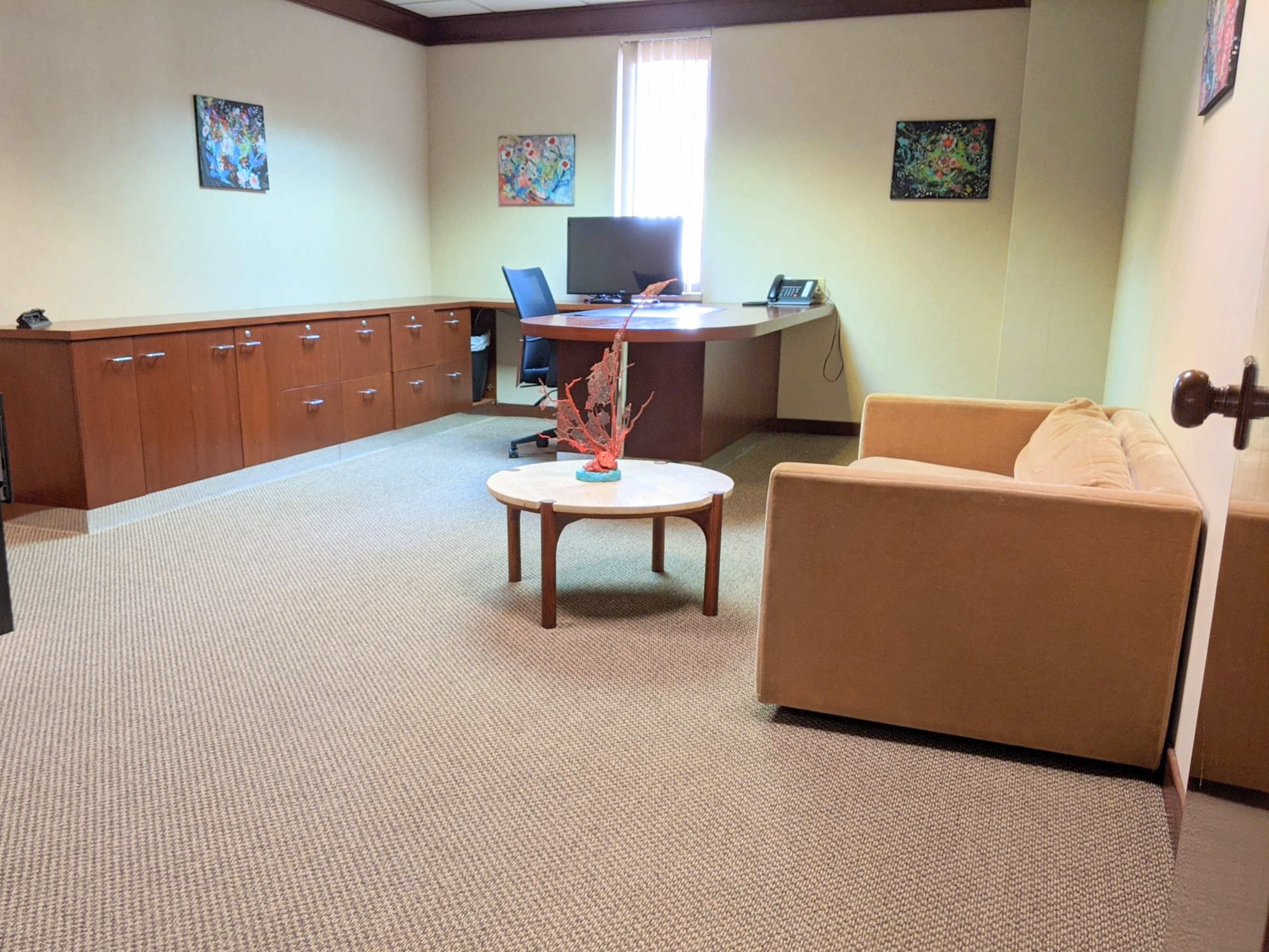 A well-lit office features a desk with a computer, a beige sofa, a round coffee table, and wooden storage cabinets along the wall.