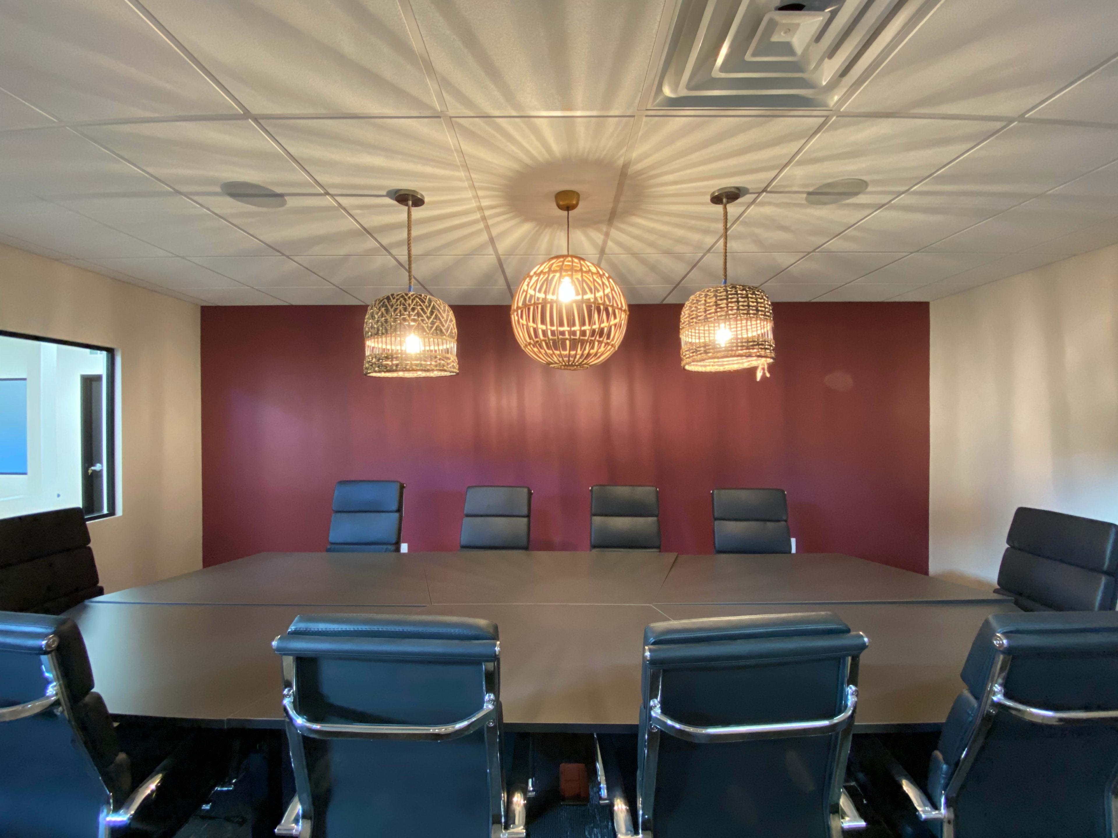 A modern conference room features a long table surrounded by black chairs, with pendant lights hanging above and a burgundy accent wall.