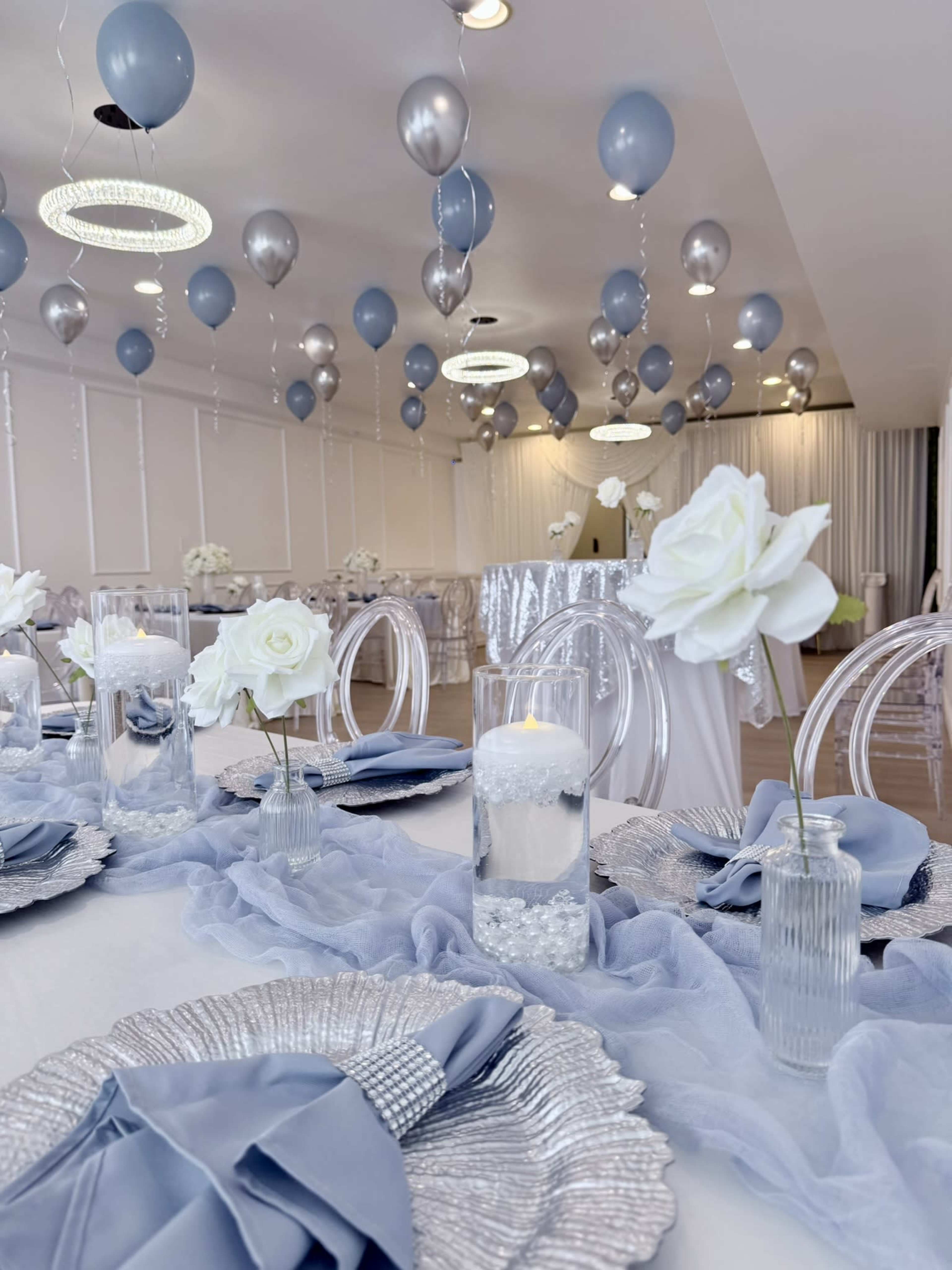 The image shows a decorated banquet room with tables set with elegant place settings, blue and silver balloons hanging from the ceiling, and light floral centerpieces.