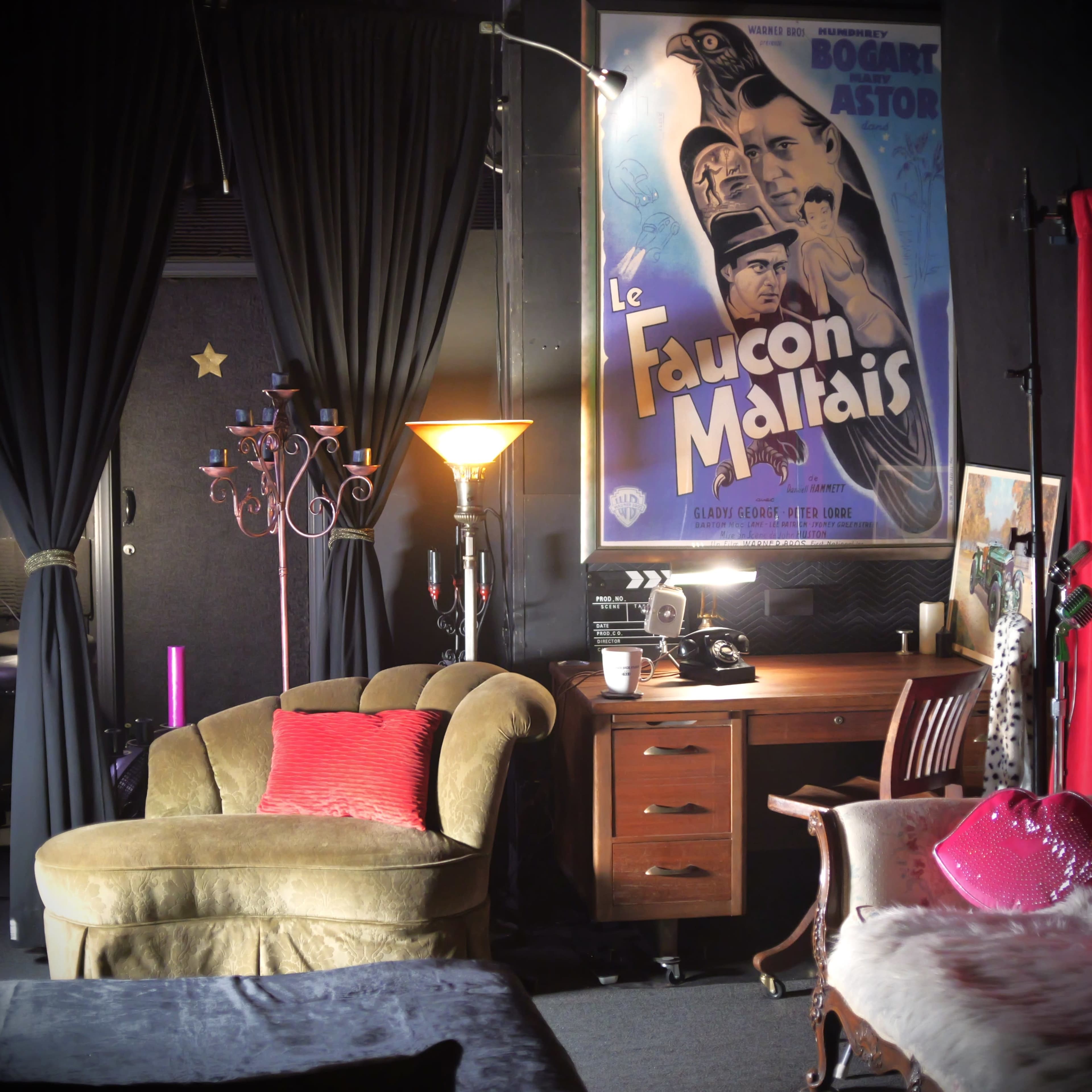 The image depicts a cozy room featuring a vintage-style sofa, a wooden desk with a typewriter, and a large film poster on the wall, surrounded by elegant decor and draped curtains.