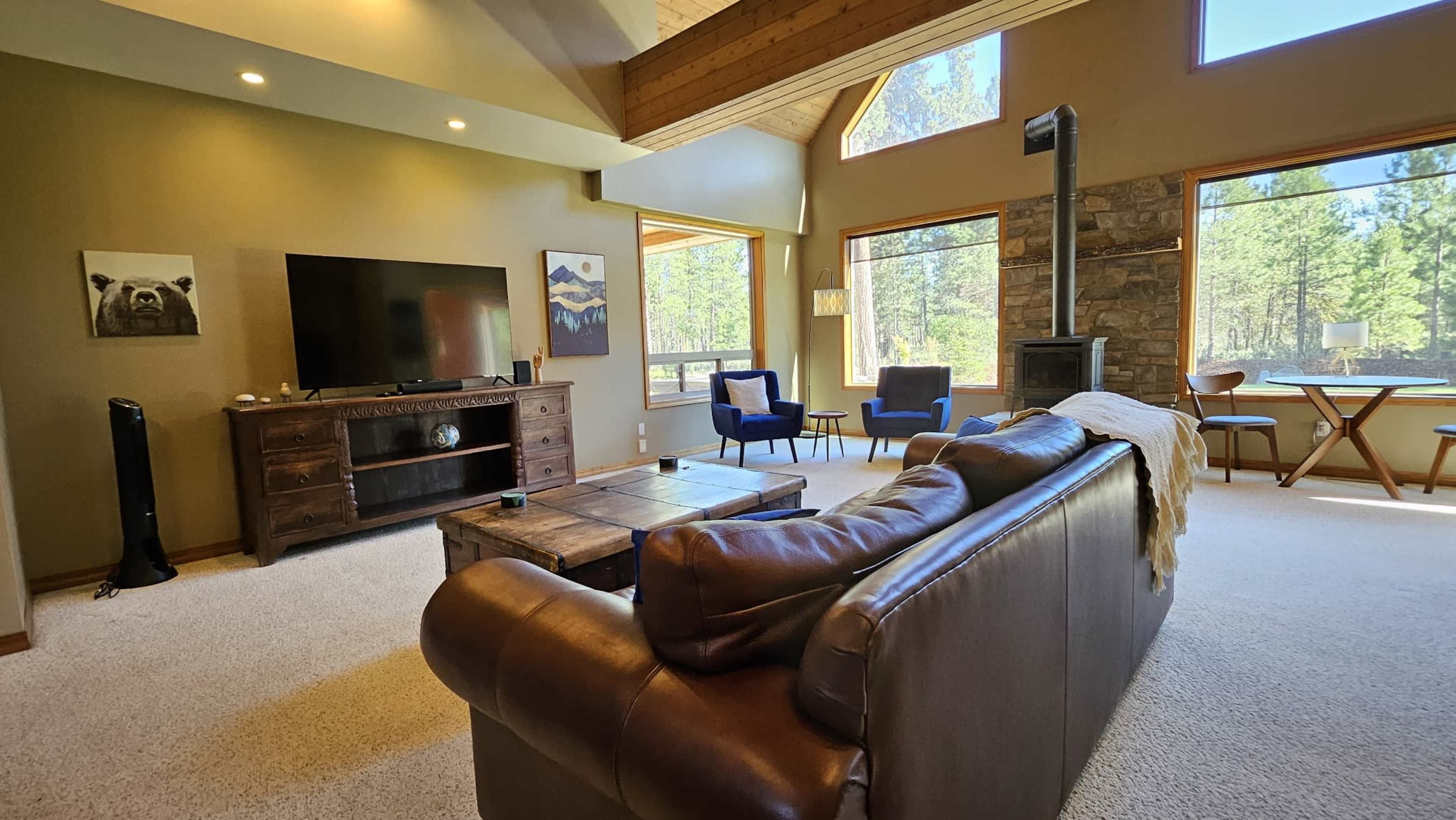 The image shows a cozy living room with a leather sofa, a wooden coffee table, a television on a cabinet, and large windows overlooking a forested area.