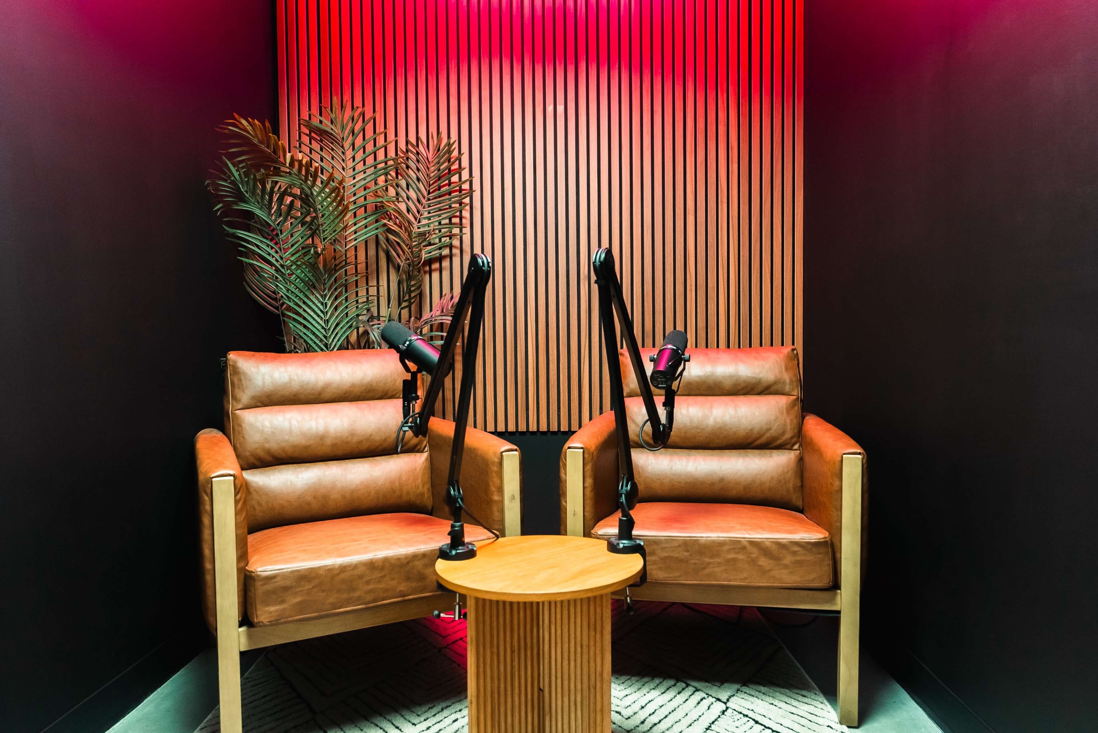 Two brown leather chairs facing each other with a circular wooden table in between, set against a backdrop of dark walls and illuminated by pink lighting.