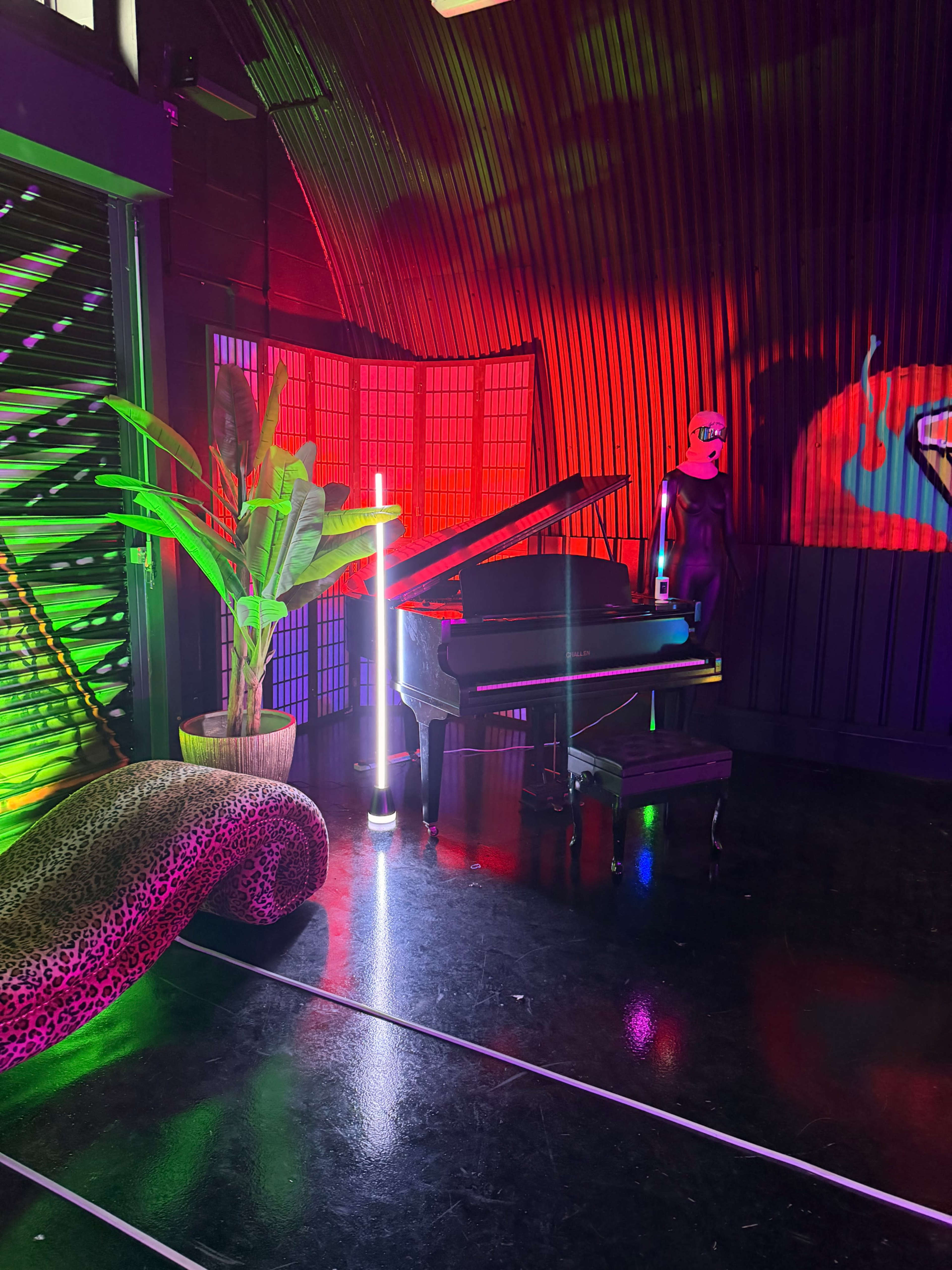 The scene features a black grand piano surrounded by vibrant neon lights, a decorative plant, and a stylized mannequin, all set against a backdrop of colorful illuminated panels.
