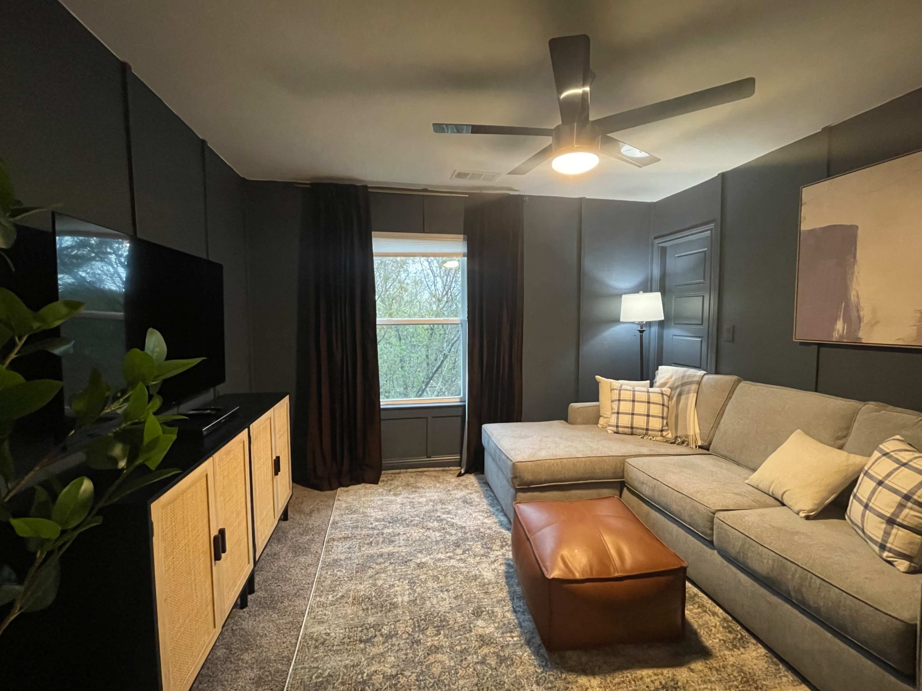 The image shows a modern living room featuring a sectional couch, a coffee table, a floor lamp, a television on a cabinet, and a window with curtains.