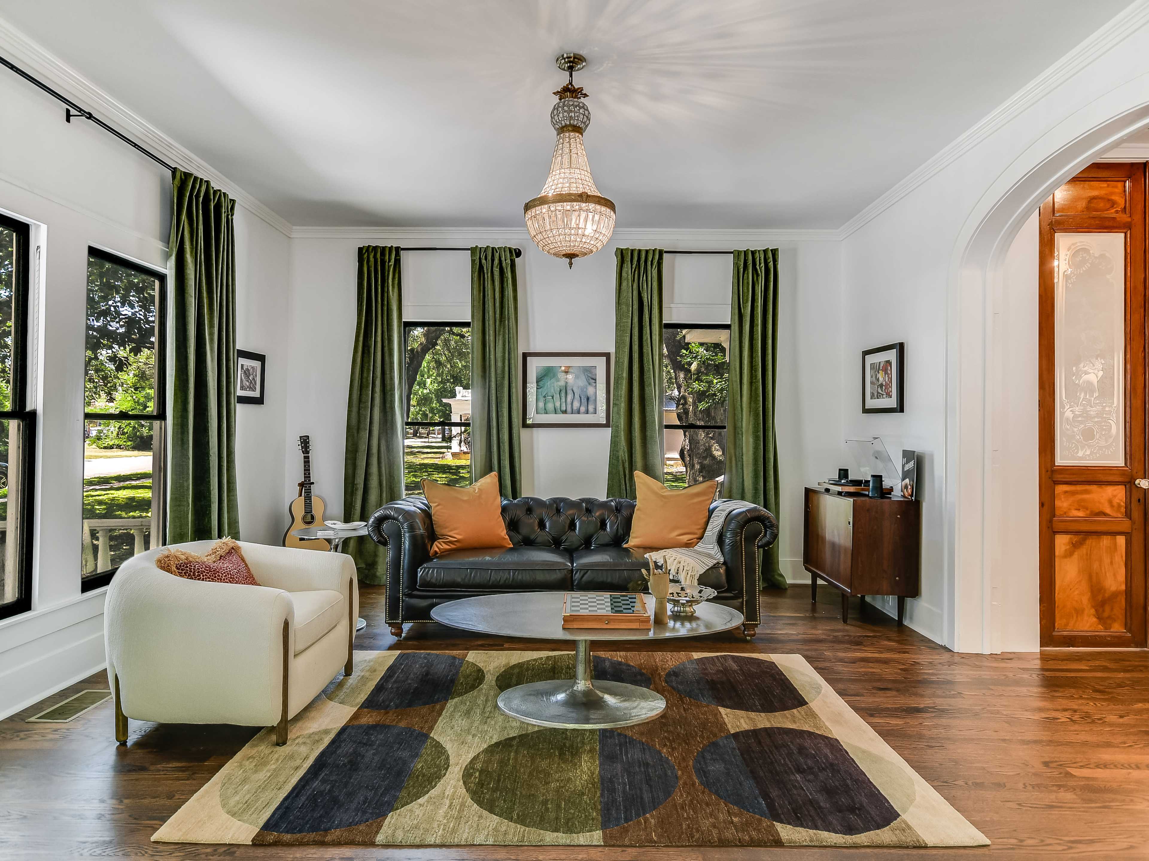 A living room features a black leather sofa, a white armchair, a circular coffee table, and large windows draped with green curtains.