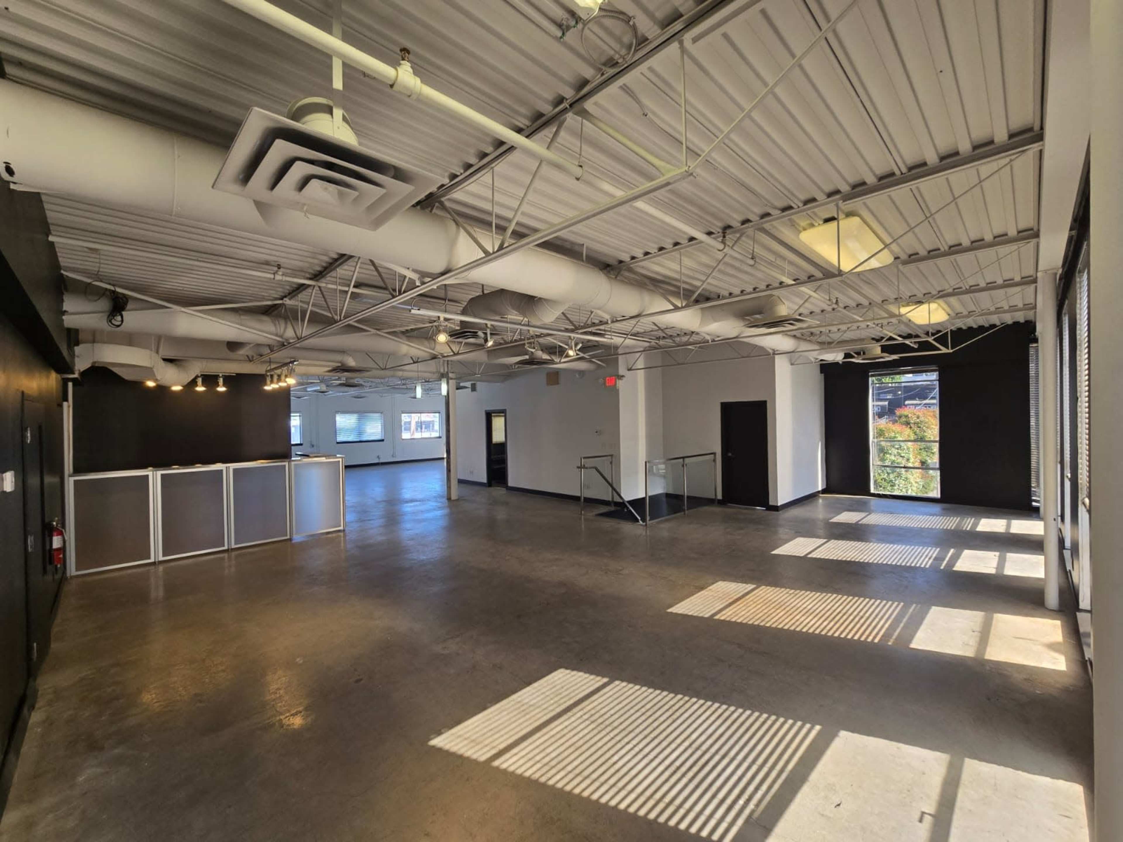 The image shows an open industrial space with exposed metal beams, large windows, and shadows cast on the floor from sunlight.