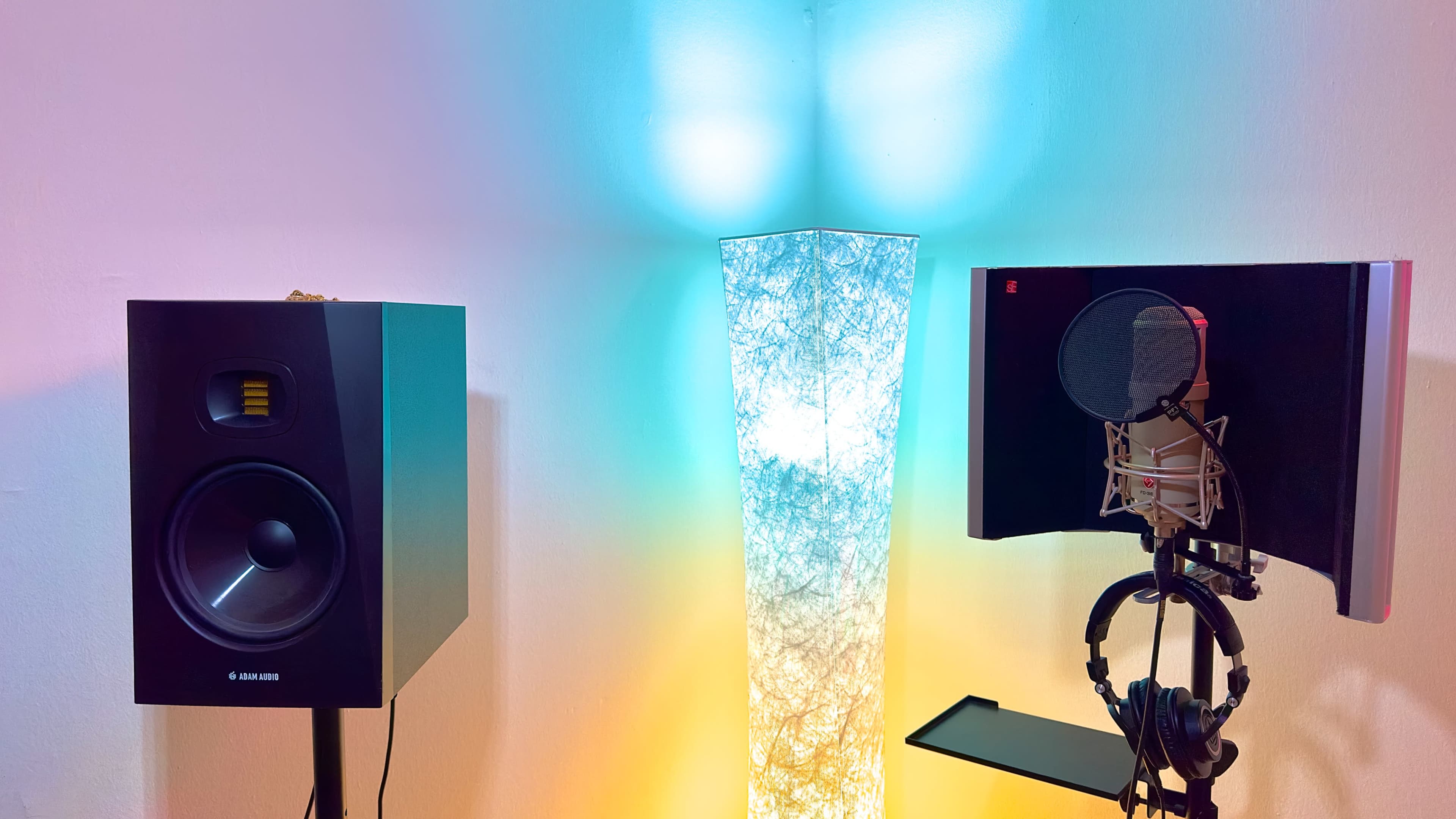 A pair of speakers flanking a microphone setup with a pop filter, situated next to a decorative lamp that emits soft colored light.