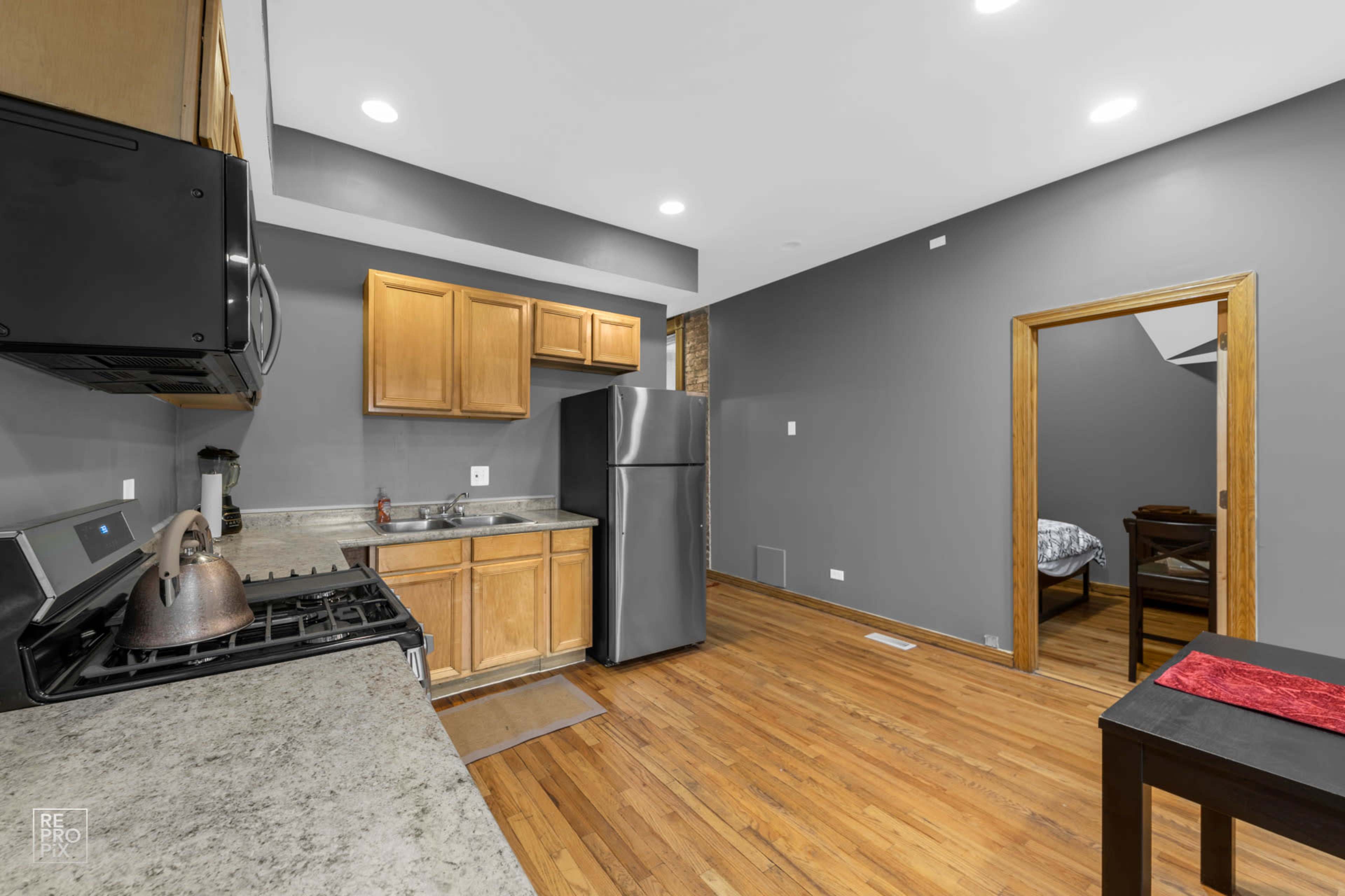 A modern kitchen with wooden cabinets, a stainless steel refrigerator, and hardwood floors, adjacent to a small dining area.