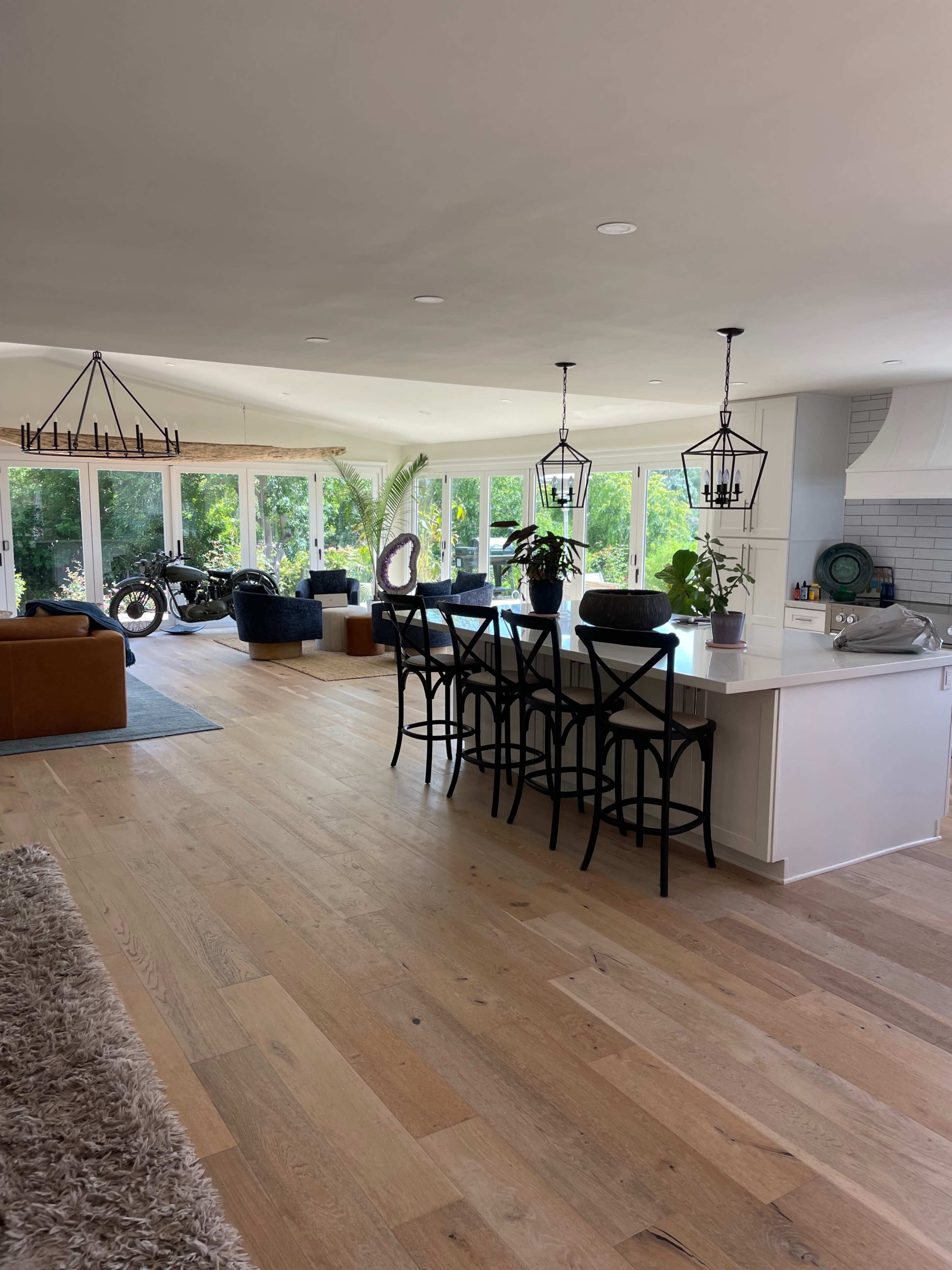 An open-concept living space featuring a kitchen with a long island and seating, a dining area, and large windows that provide natural light and views of greenery outside.