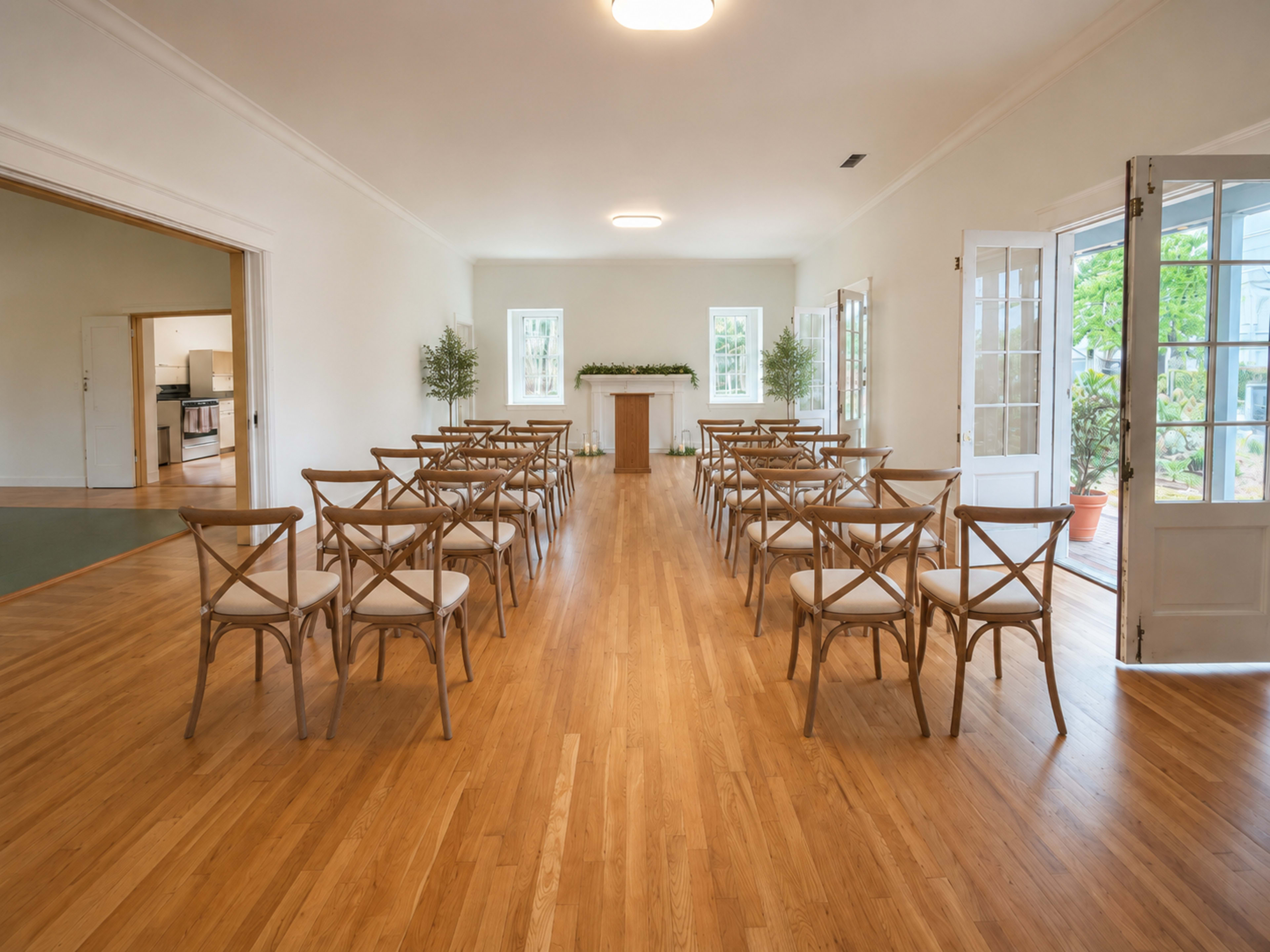The image depicts a simple, well-lit room arranged with rows of wooden chairs facing a small altar or table, with doors leading to an outdoor area.