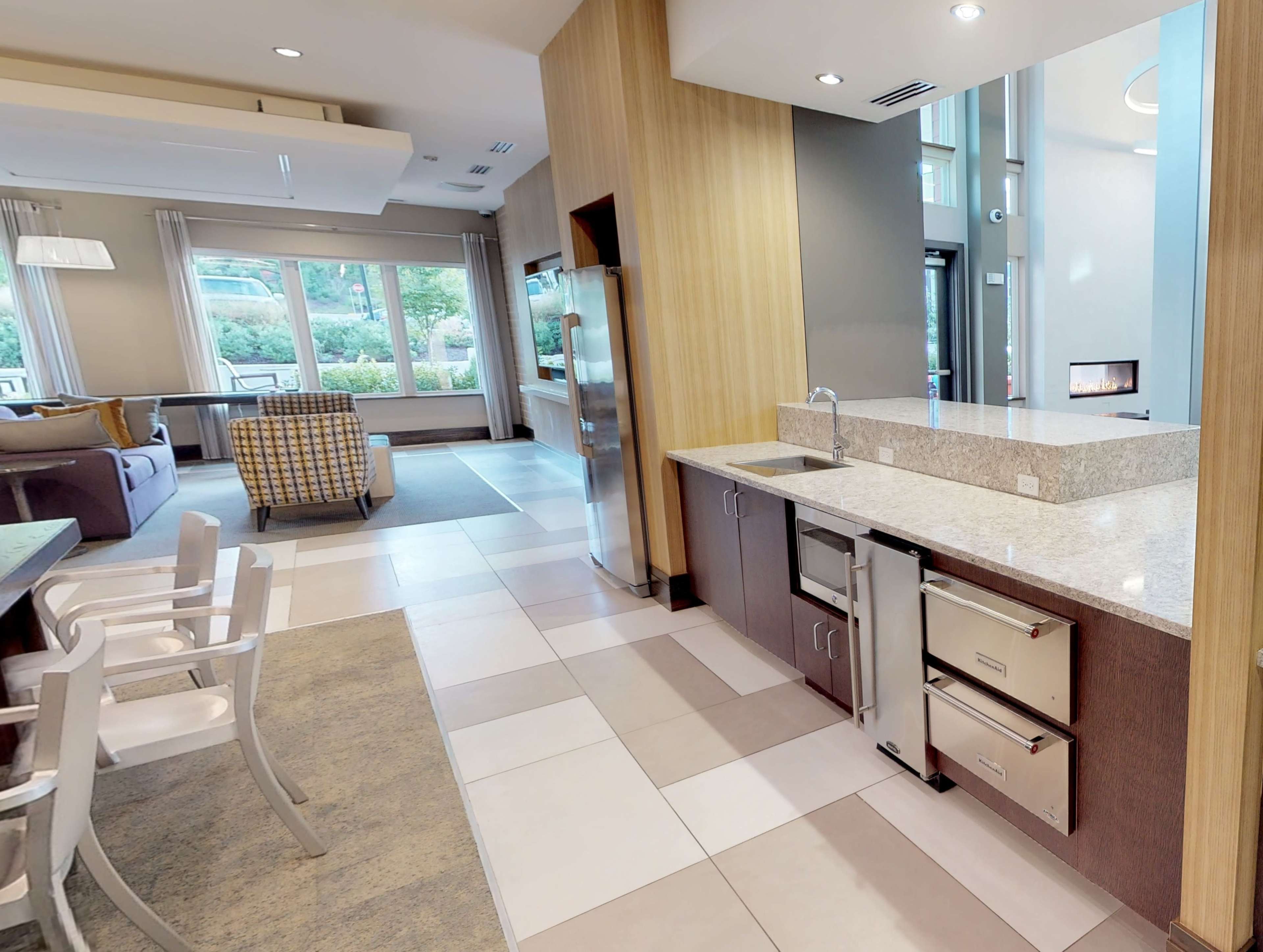 The image shows a modern living space featuring a kitchen area with a granite countertop, stainless steel appliances, and a living room furnished with a sofa and a patterned chair.