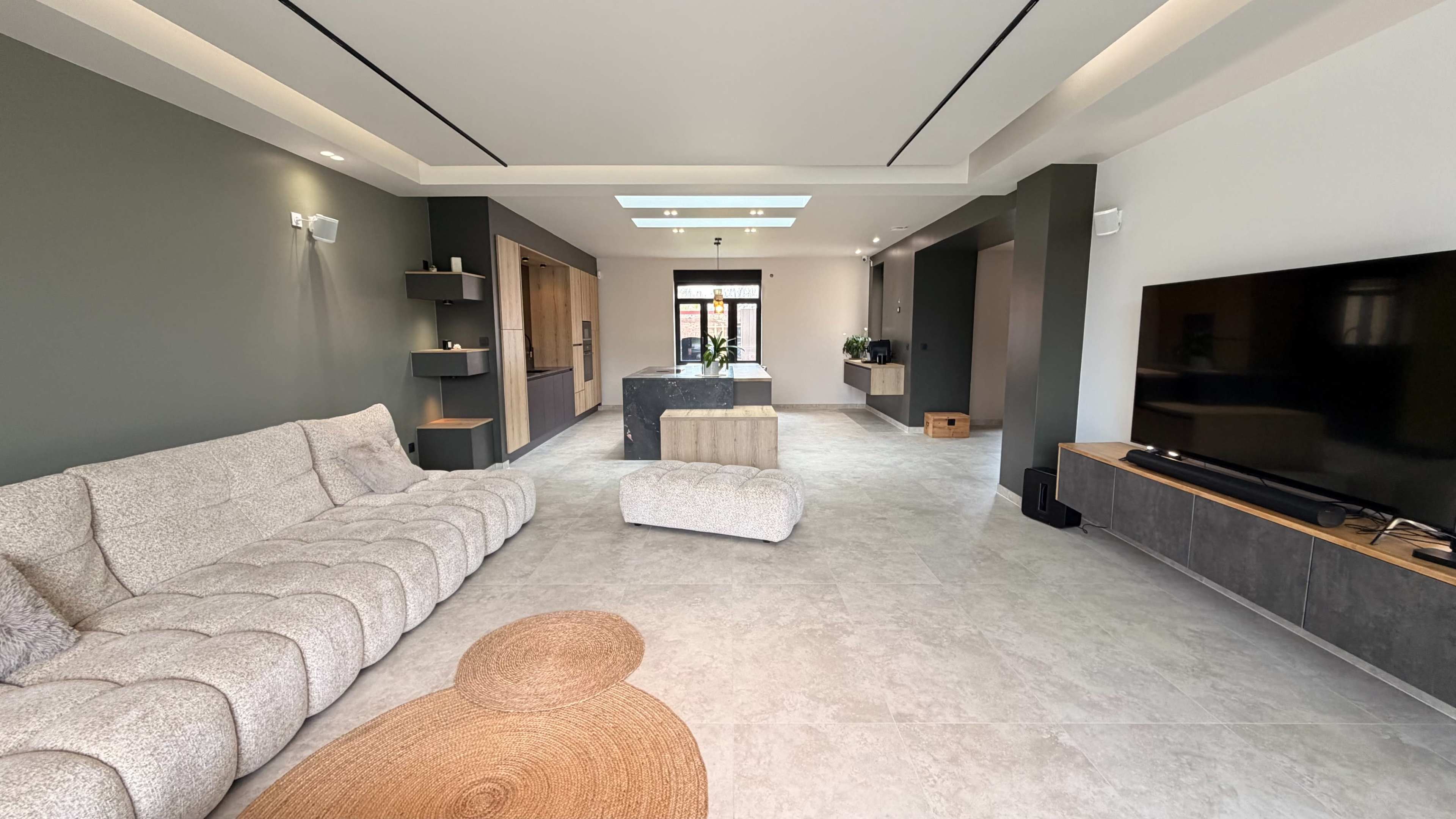 The image shows a modern living room with a light-colored sectional sofa, a large flat-screen TV on a low cabinet, and a central coffee table, featuring large windows that allow natural light to enter the space.