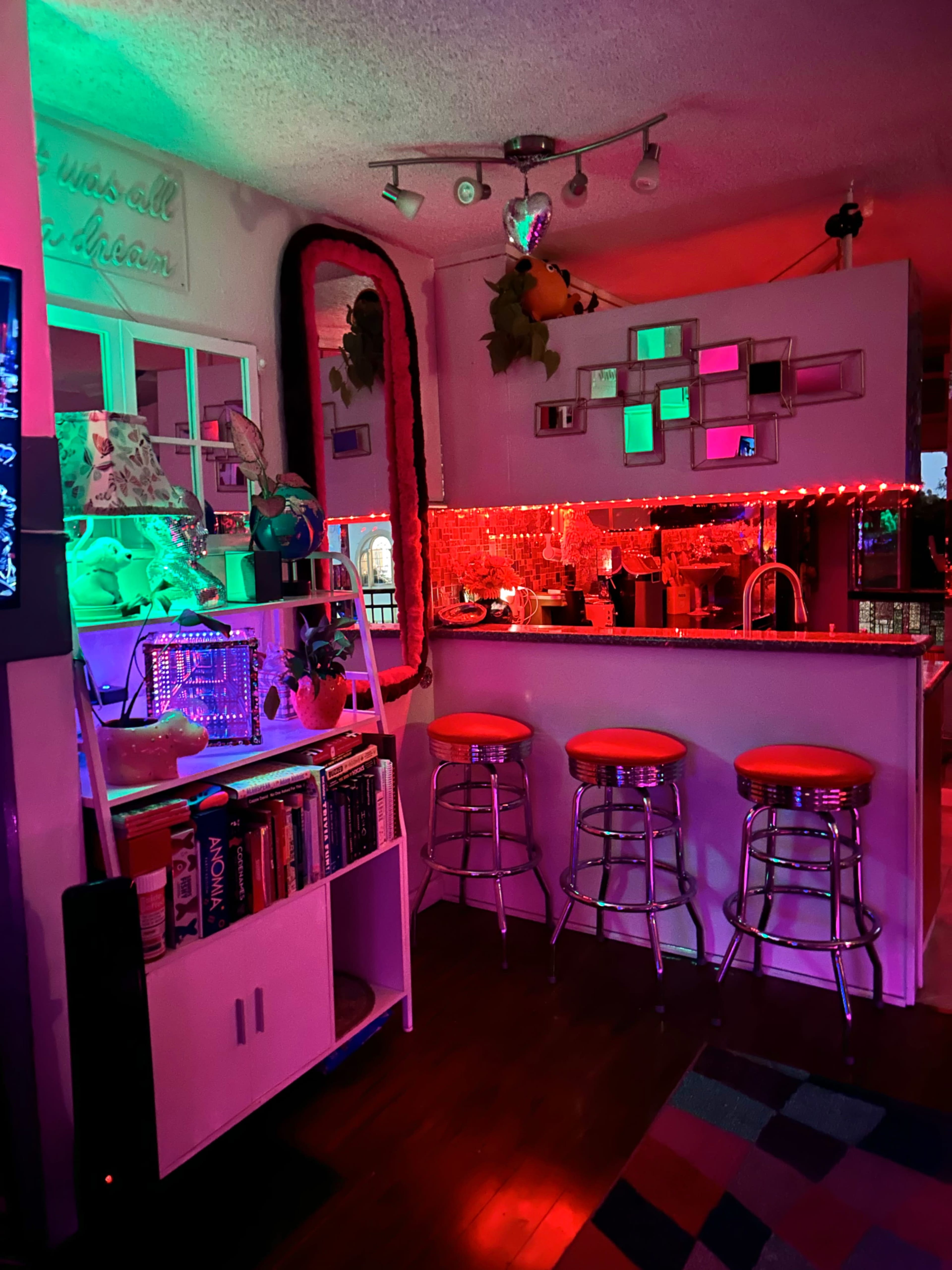 The image shows a colorful, dimly lit kitchen area with neon lights, a large mirror, and four bar stools along a countertop.