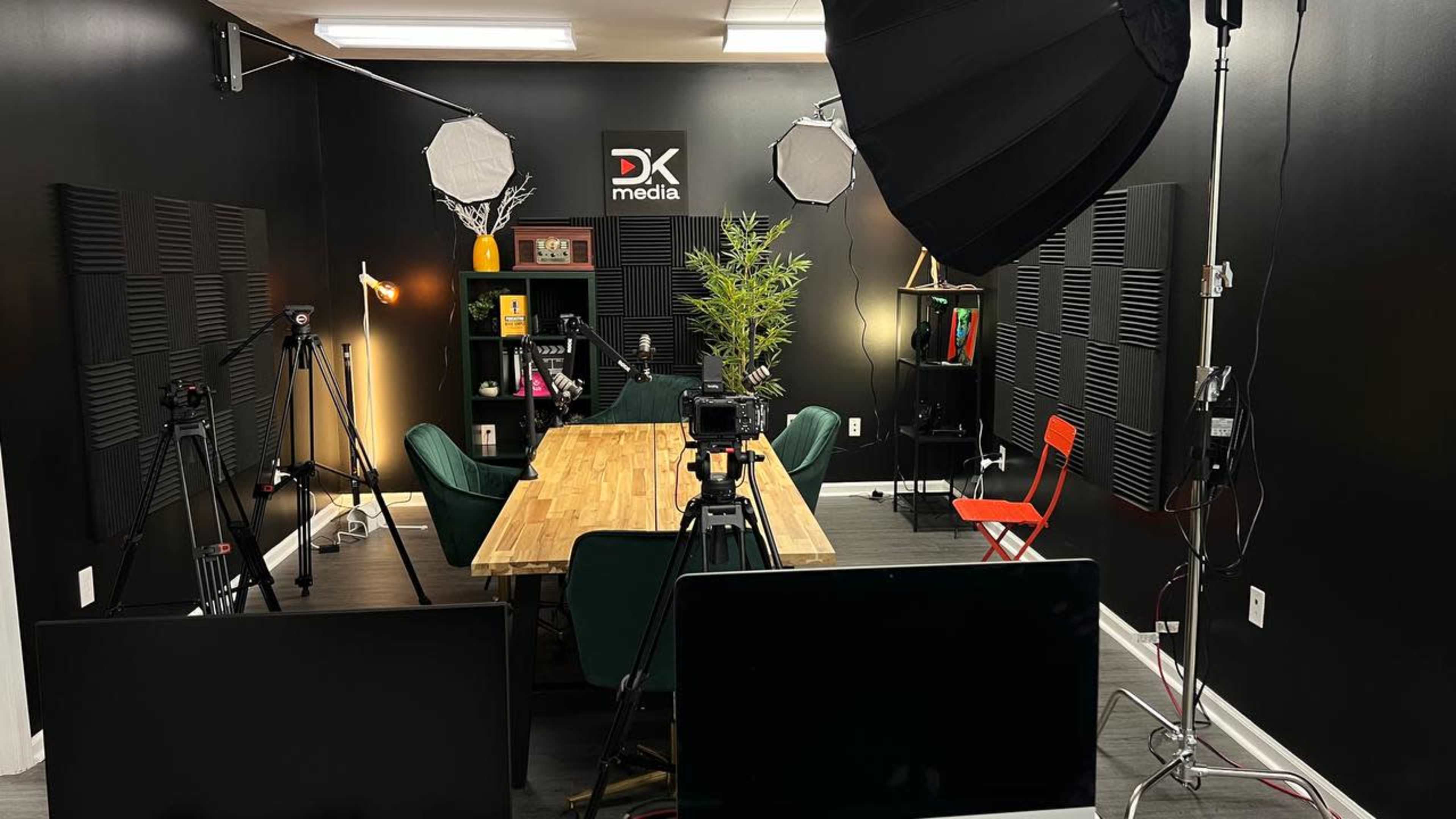 The image shows a video production studio with black walls, a large wooden table surrounded by green chairs, and various cameras and lighting equipment set up for a shoot.