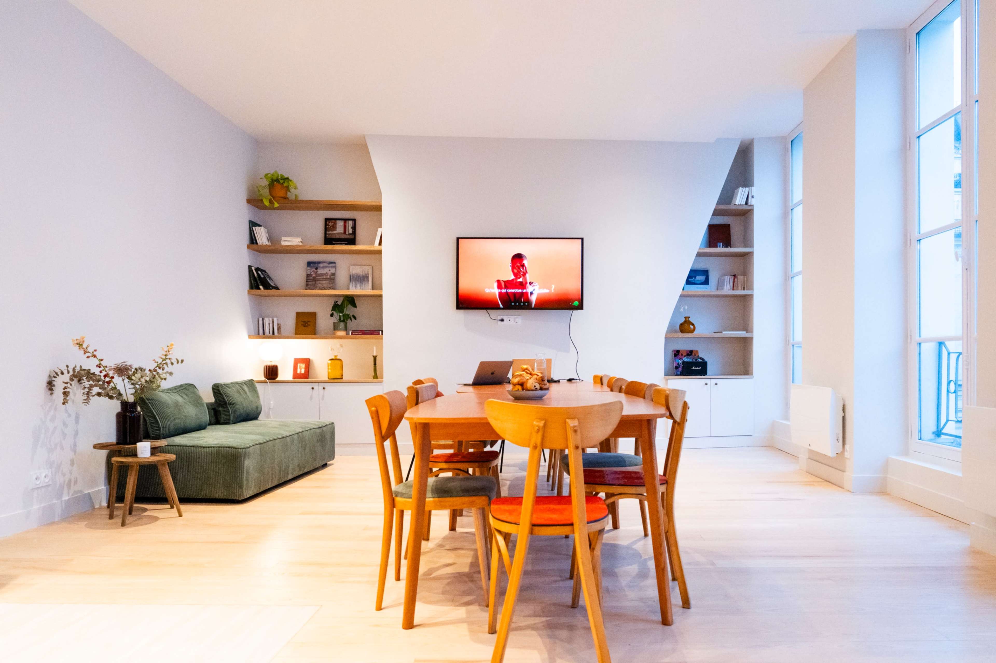 The image shows a modern living space featuring a green sofa, a dining table with wooden chairs, a wall-mounted television, and shelves with decorative items.