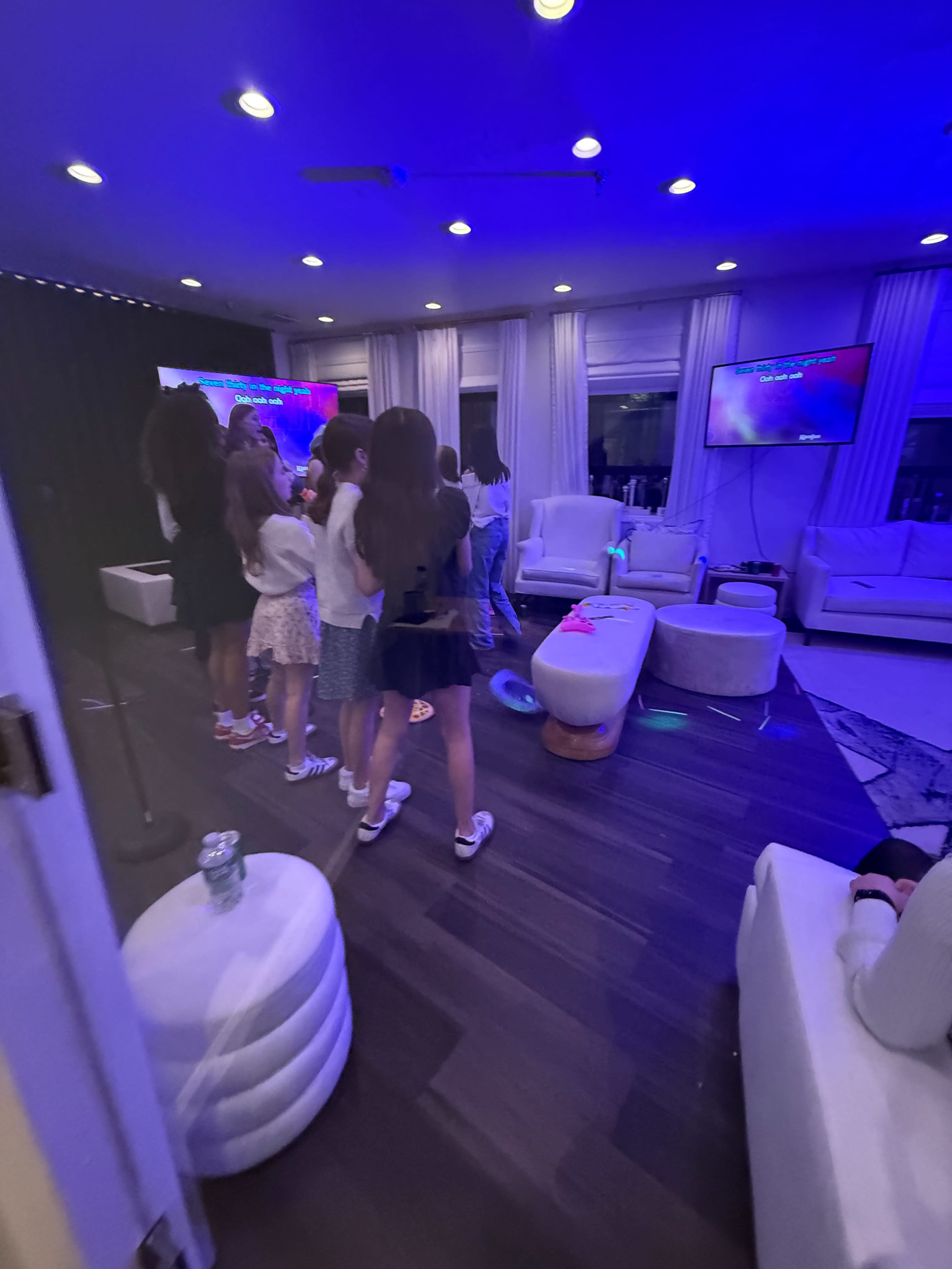A group of children is gathered in a brightly lit lounge area with blue lighting, watching a screen while standing near white furniture.
