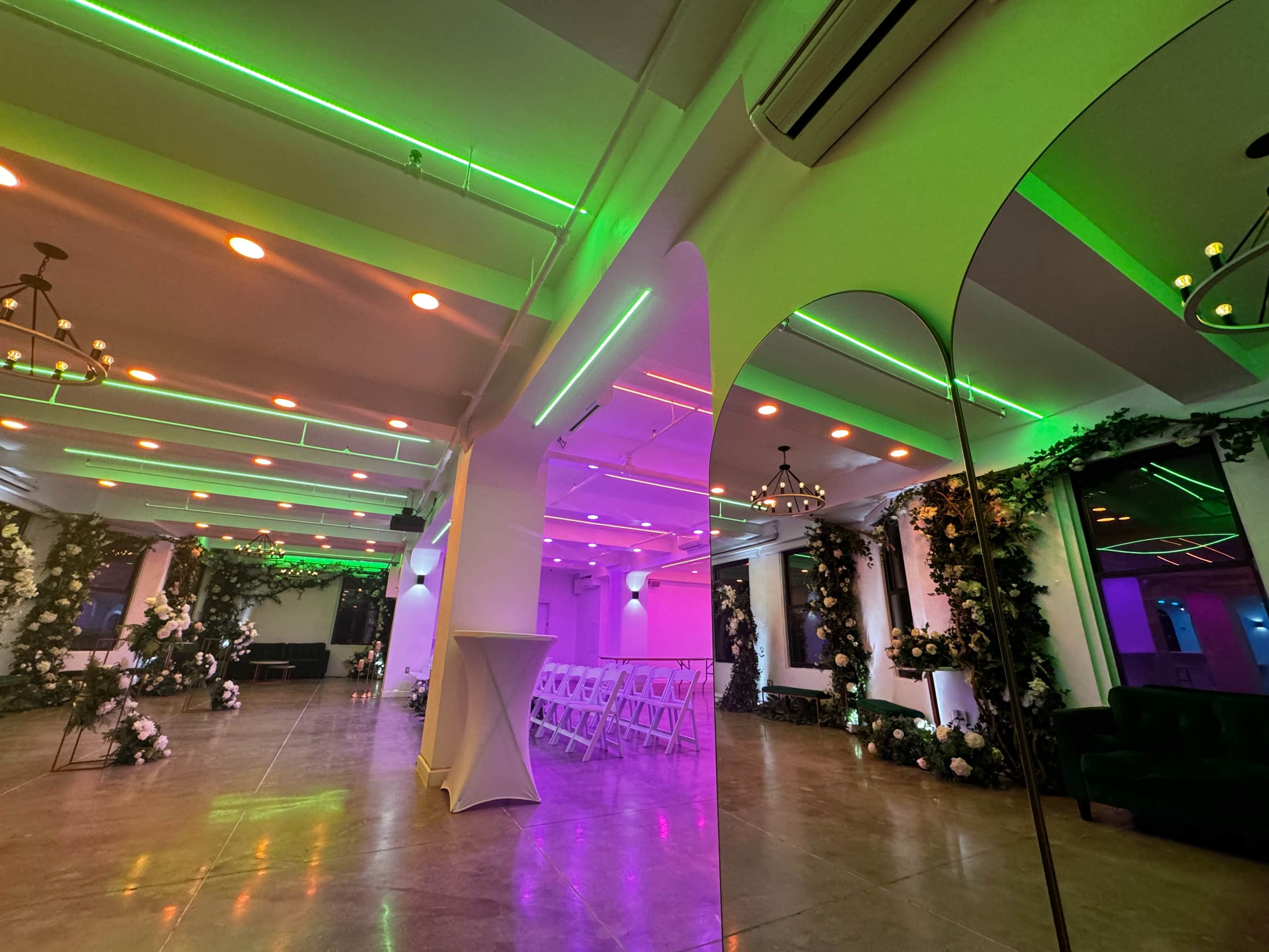 A modern event space with colorful neon lighting, floral decorations along the walls, and rows of white chairs set up for an event.