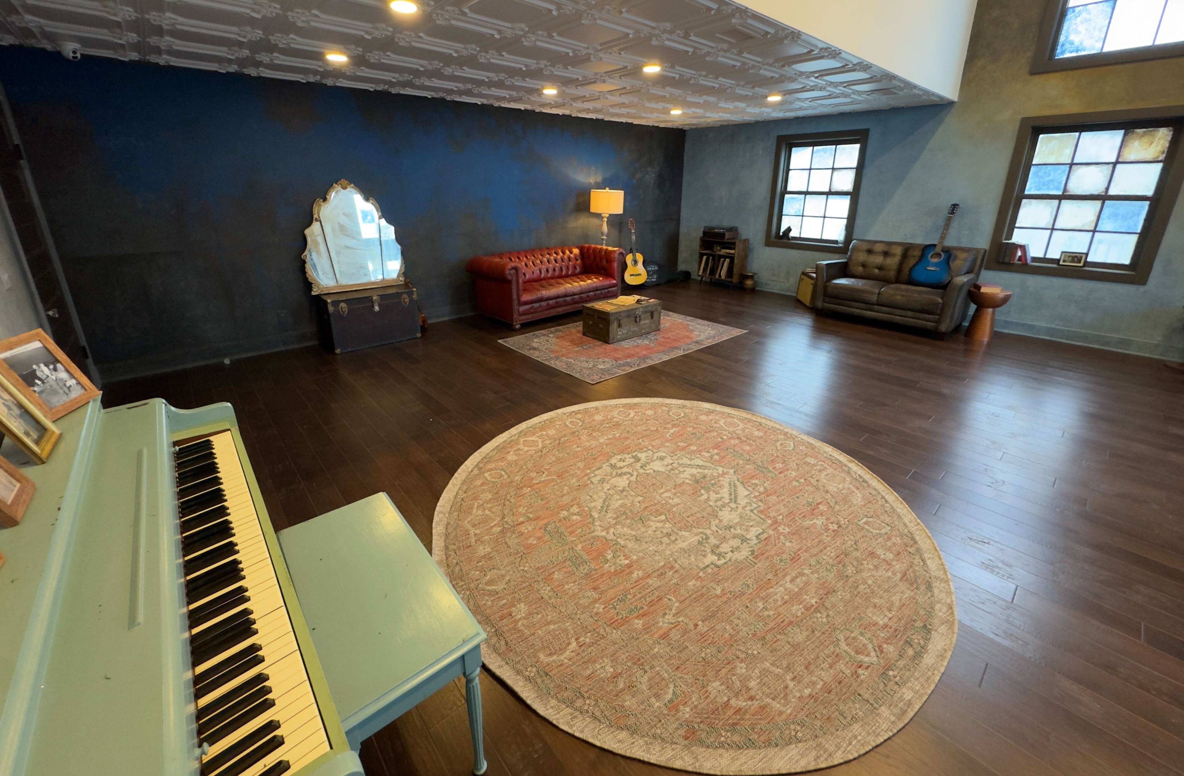 The image shows a spacious living area with a patterned rug, a green piano, a leather couch, a red sofa, and several guitars, illuminated by overhead lights.