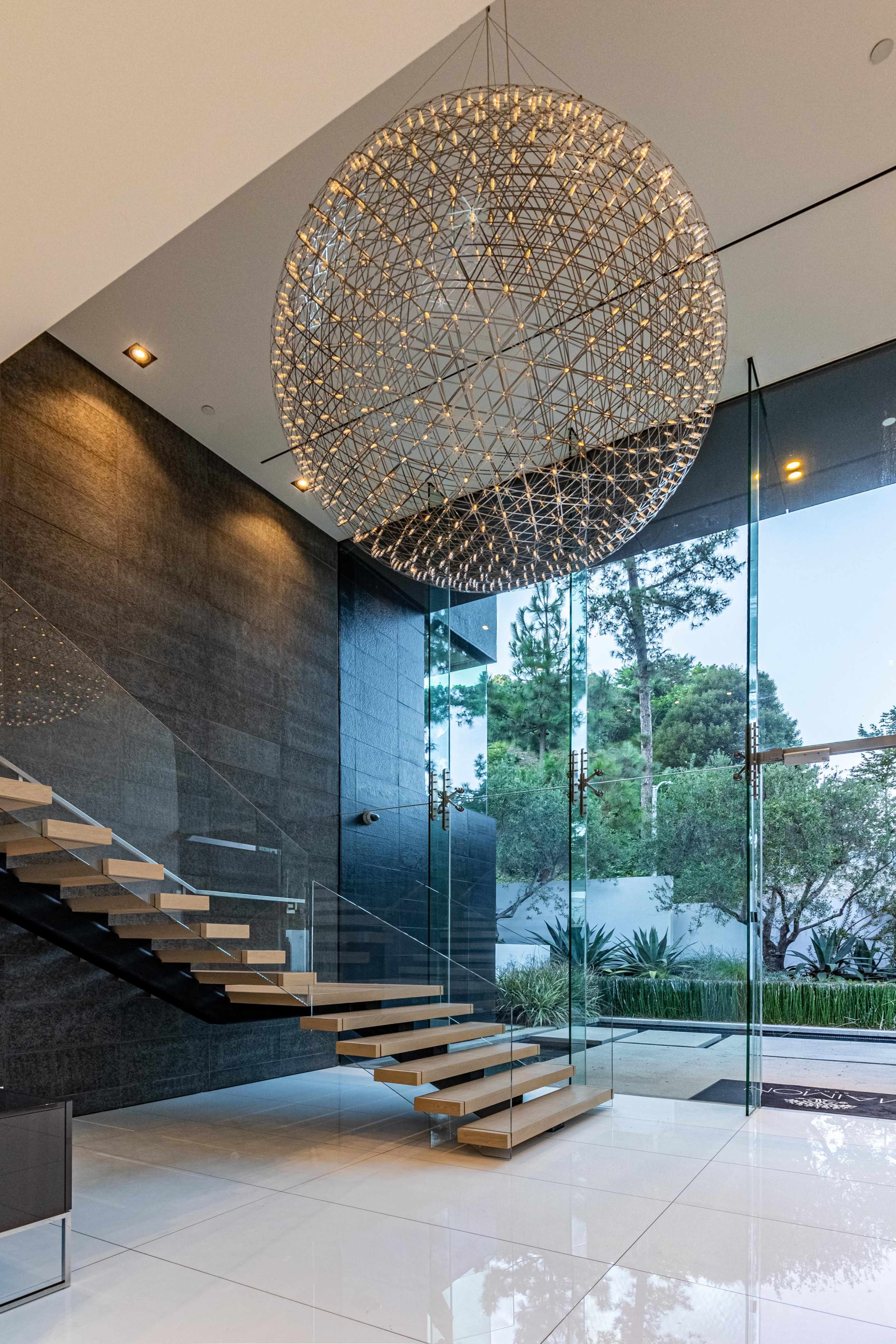 The image shows a modern entryway with a floating staircase, large glass panels, and a circular chandelier.