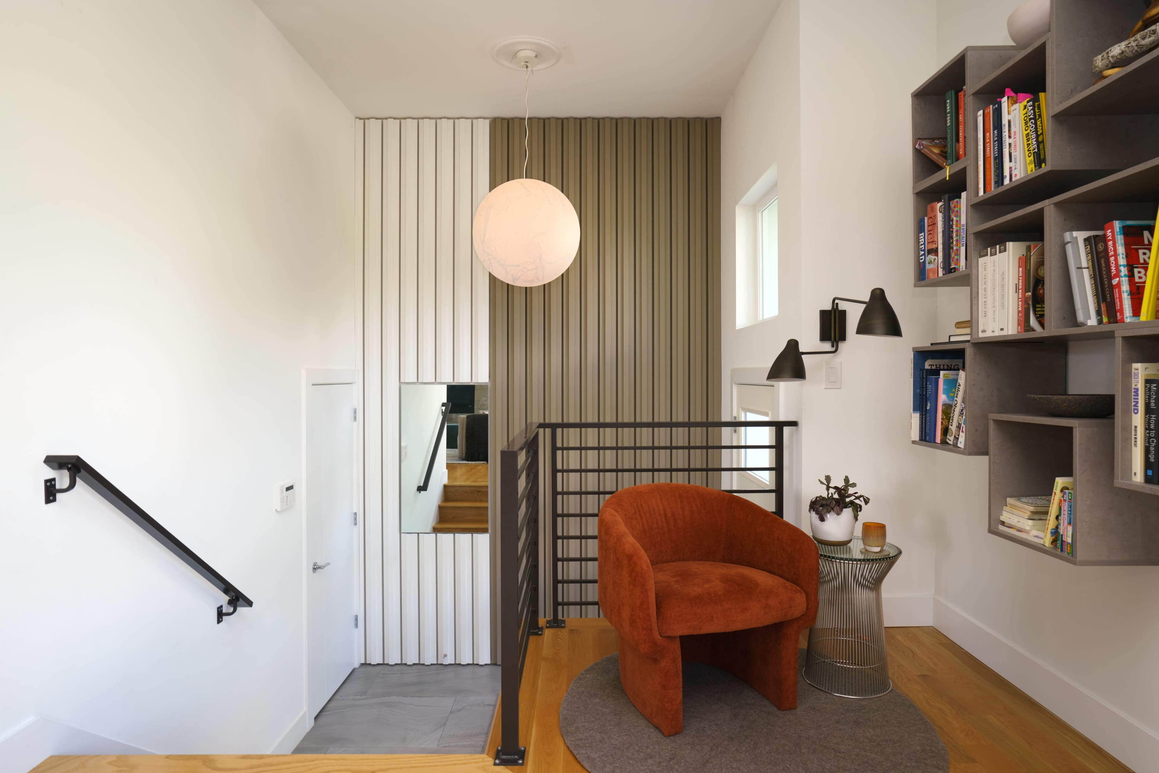 A modern entryway features a circular orange chair, a round lamp, bookshelves, and a staircase leading to an upper level.