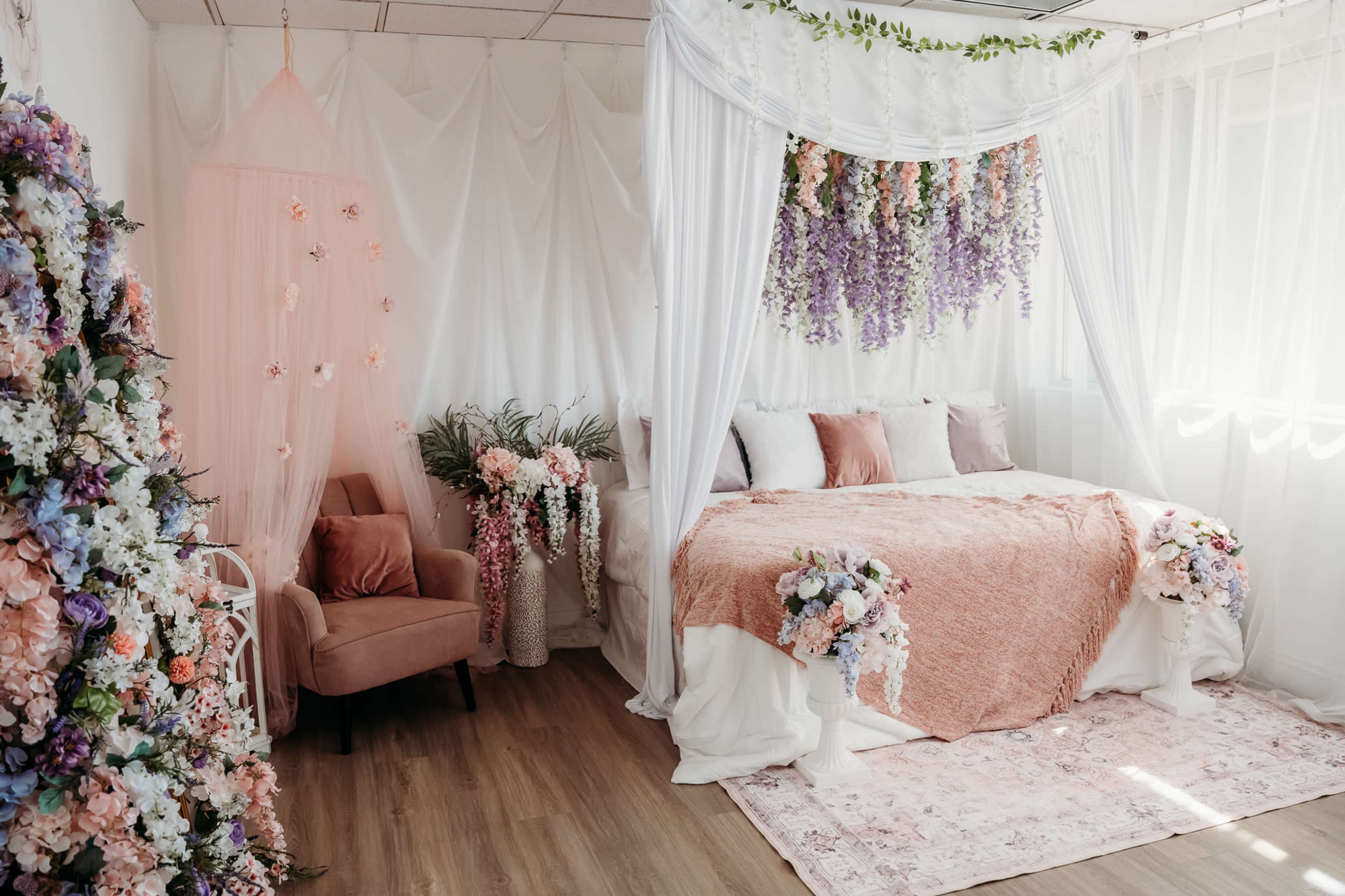 The image shows a softly decorated bedroom featuring a bed with a floral canopy, surrounded by decorative flowers, a pink armchair, and a light-colored rug.
