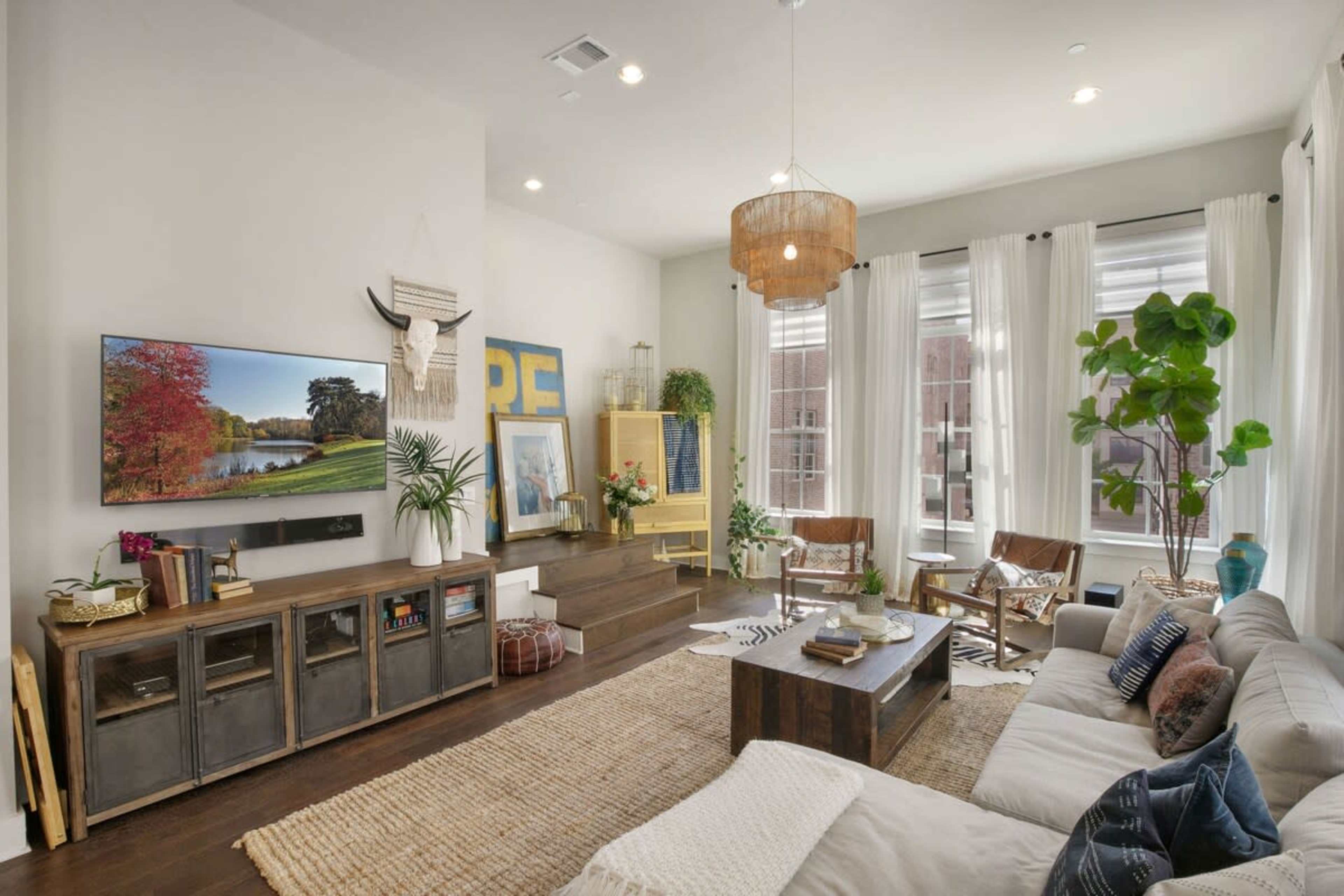 The living room features a flat-screen TV on the wall, a large coffee table, and a mix of seating including a sectional sofa and wooden chairs, all arranged around a neutral-colored area rug.