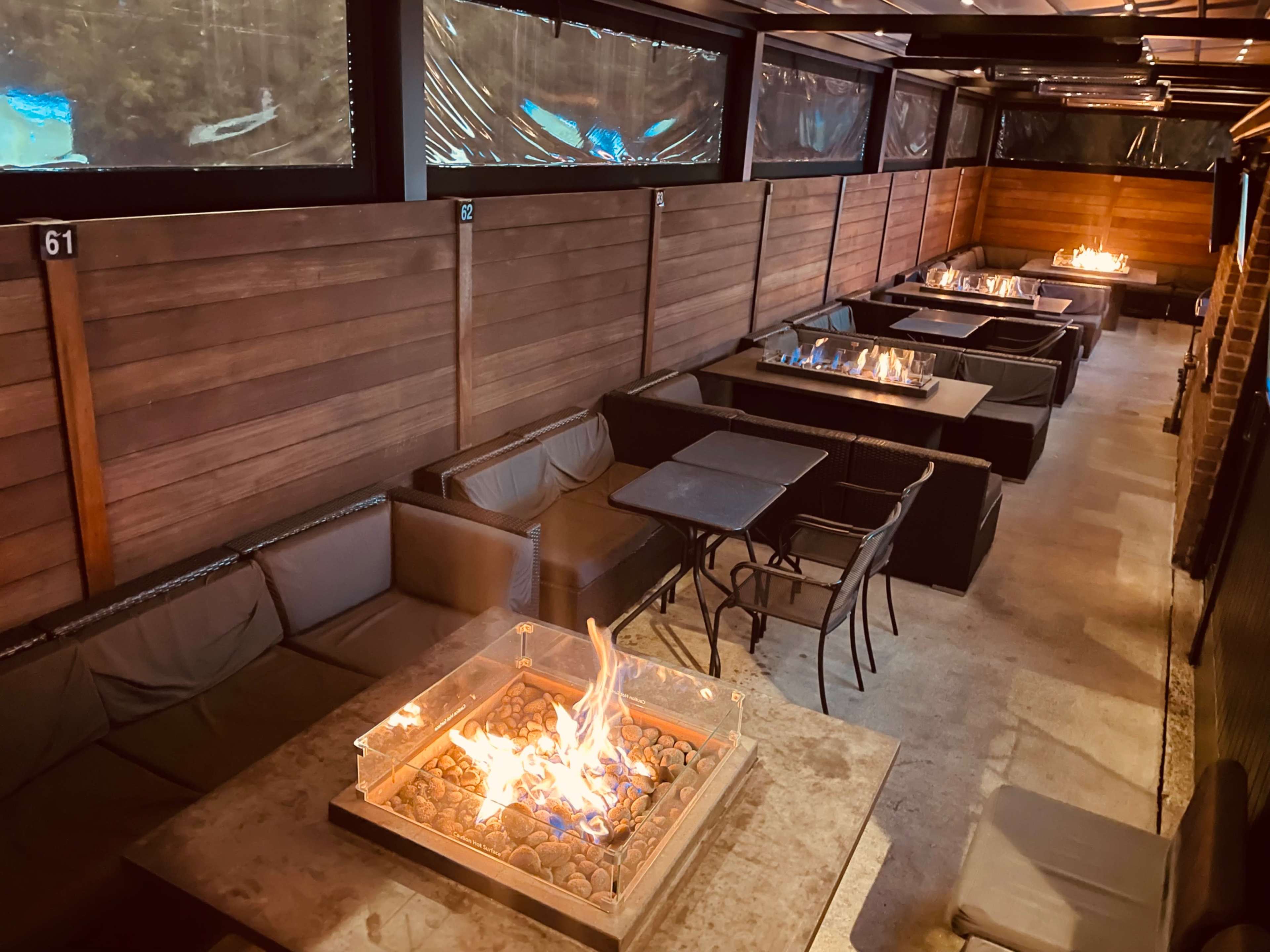 The image shows a modern outdoor lounge with several tables featuring fire pits, surrounded by wooden walls and a ceiling.