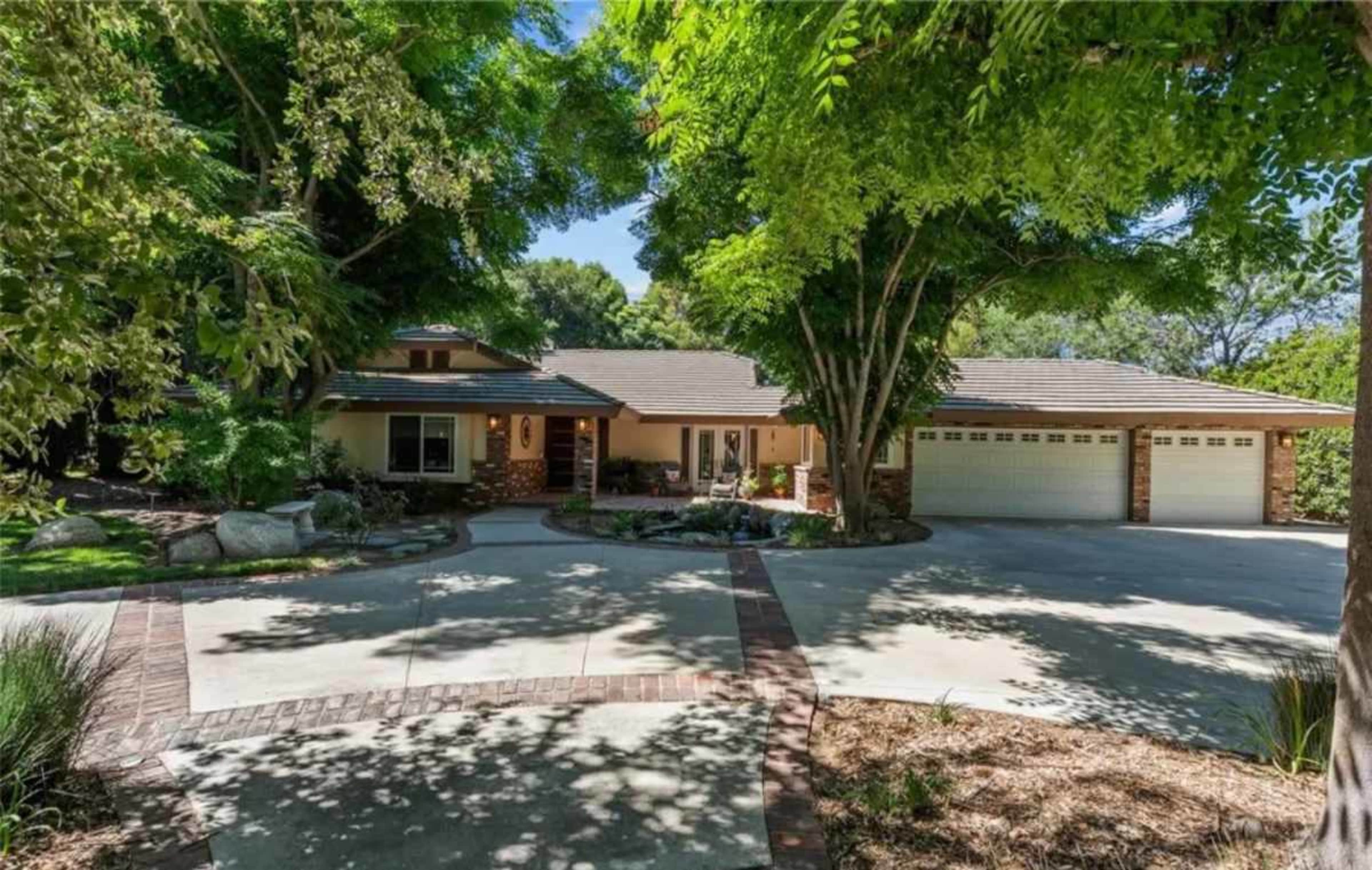 A single-story home with a three-car garage is set back from the driveway, surrounded by trees and landscaped greenery.