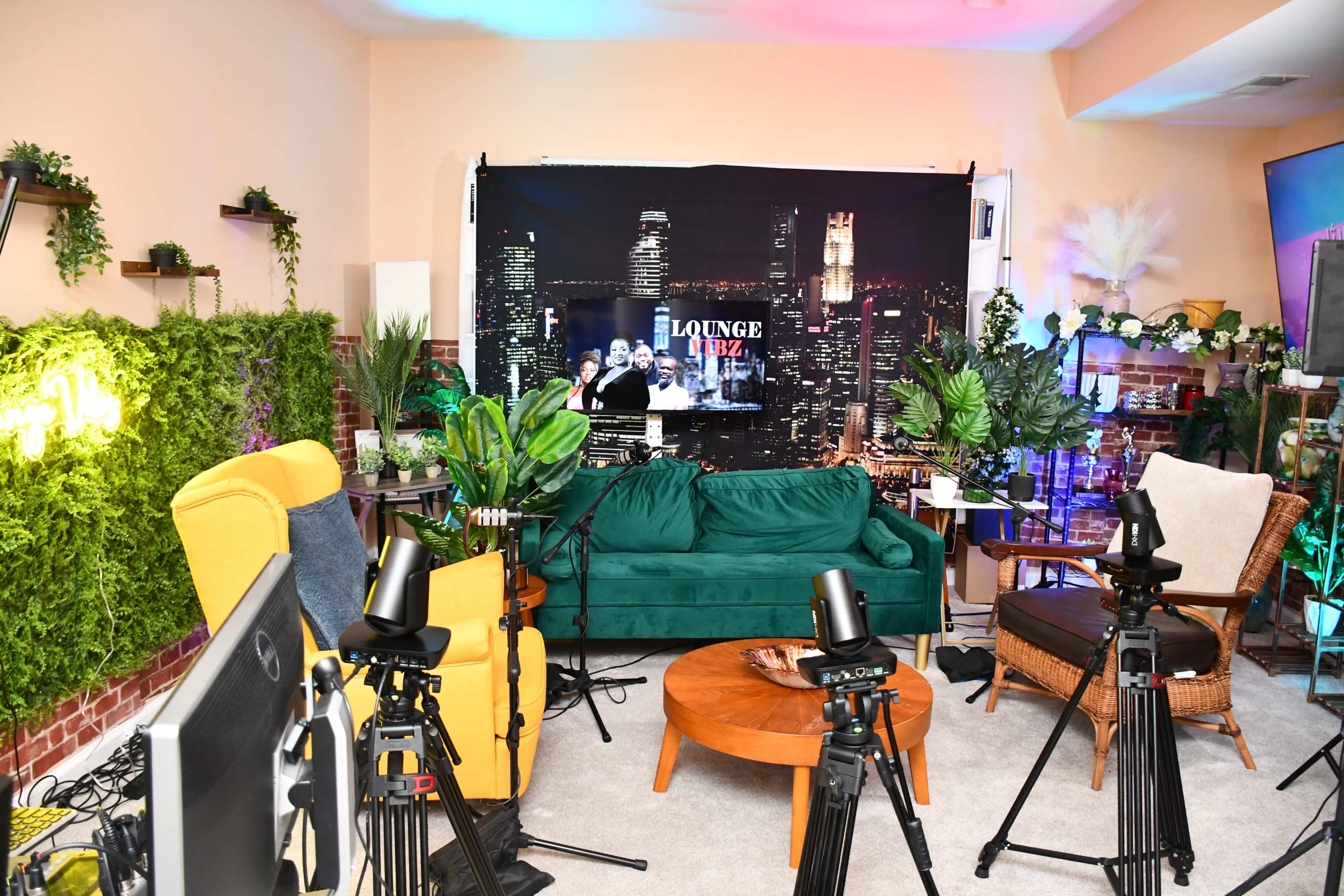 The image shows a creatively designed indoor setting with a green couch, colorful chairs, a wooden coffee table, and several cameras aimed at a backdrop featuring a city skyline.