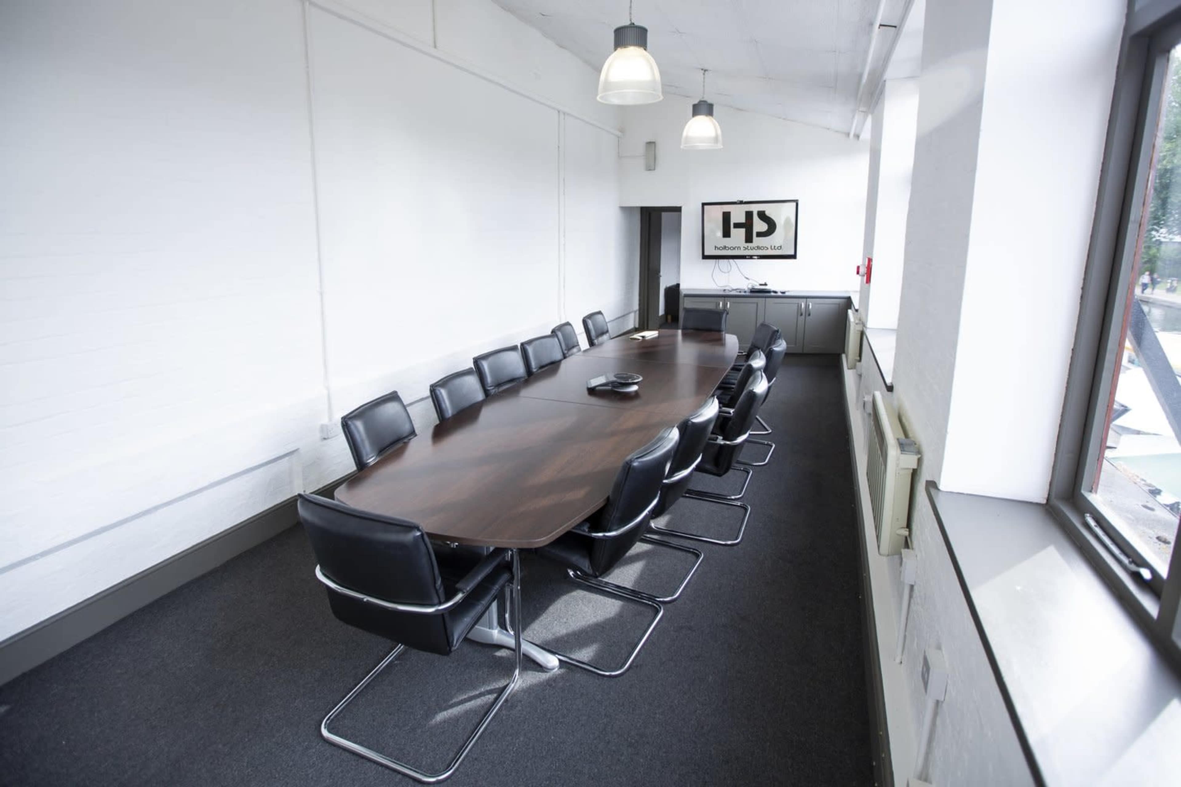 A long conference table with black chairs sits in a bright, modern meeting room featuring large windows and a wall-mounted screen.
