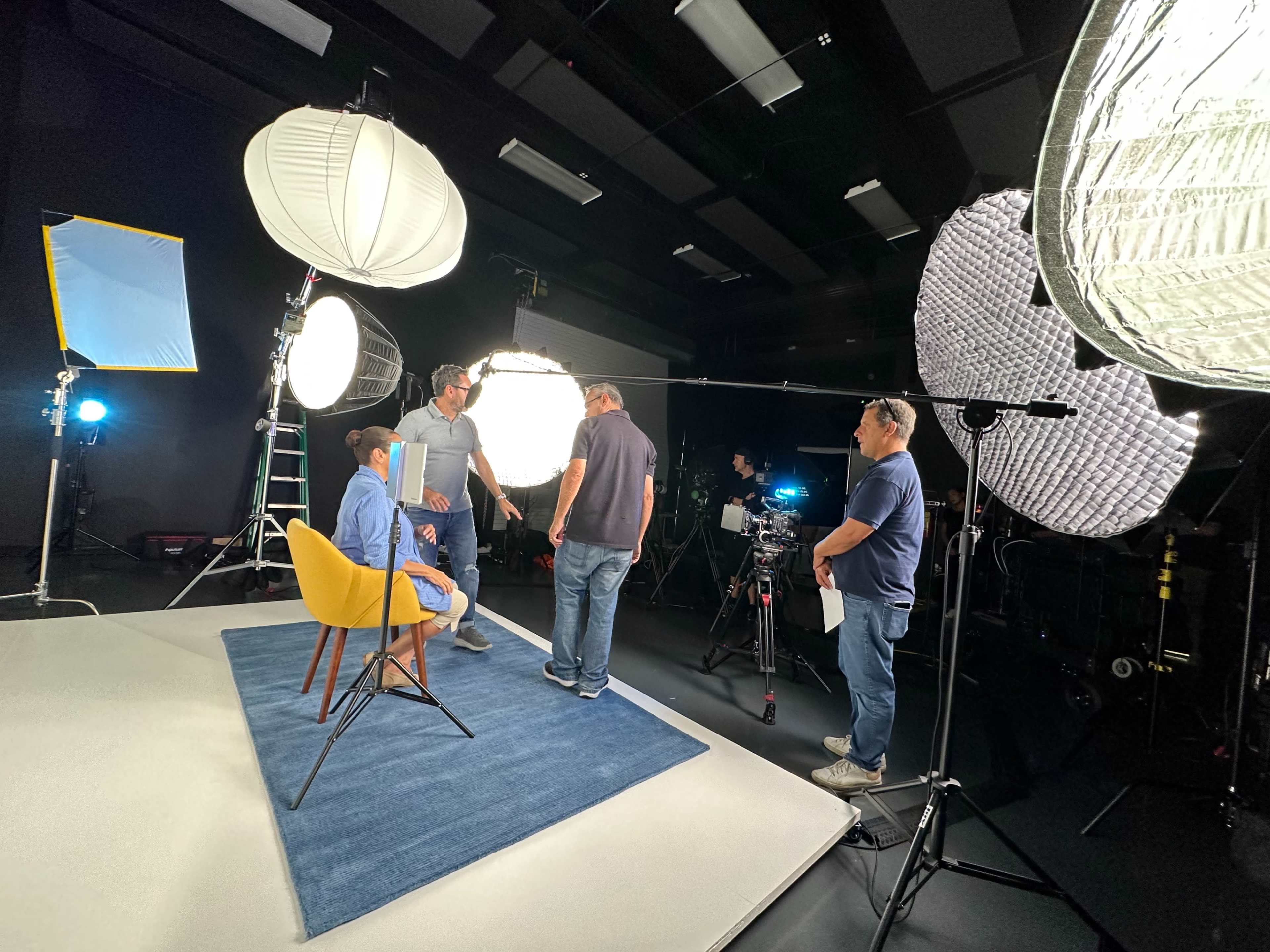 A film crew sets up lighting and equipment around a seated individual in a studio.