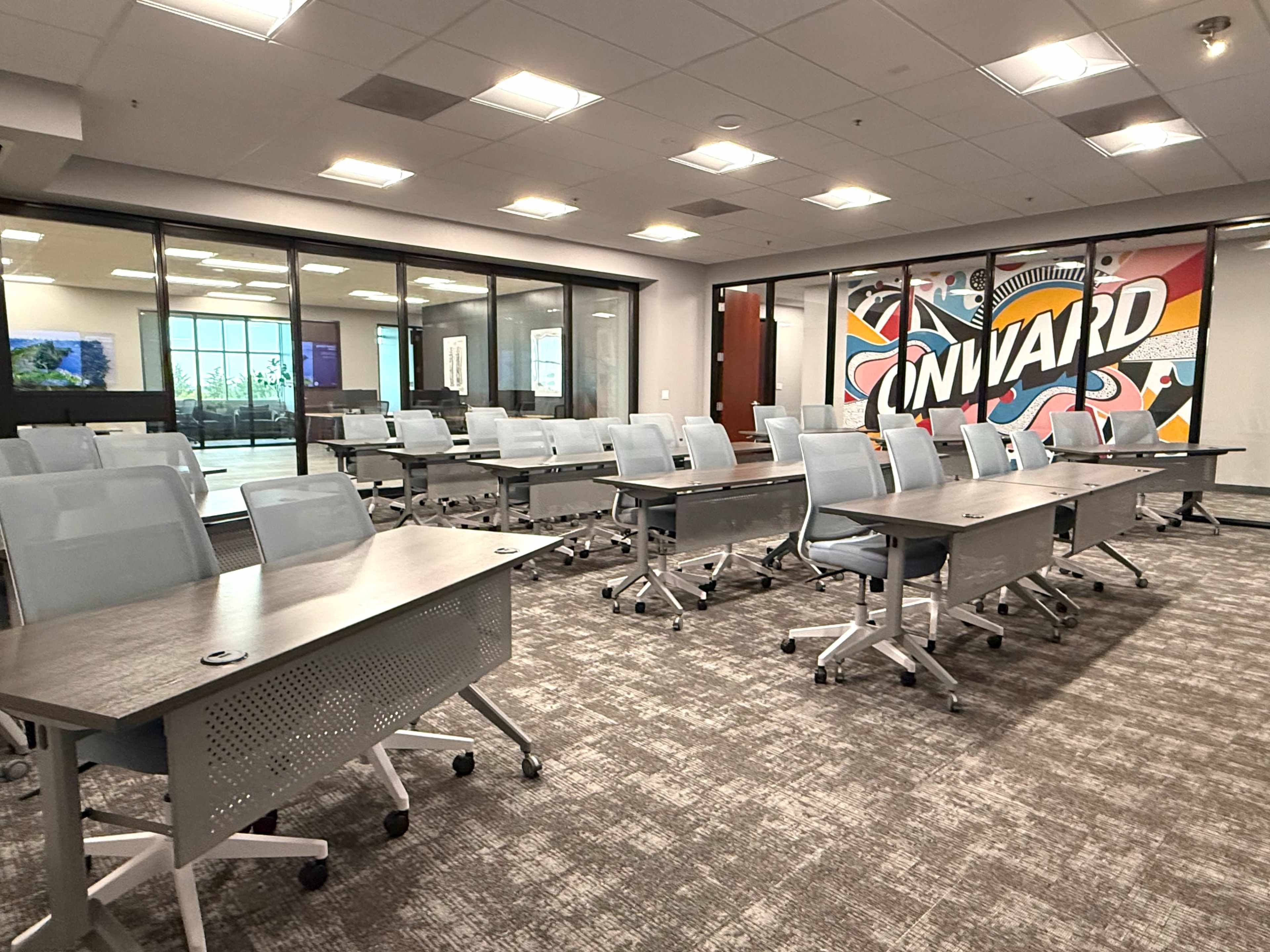 The image shows a modern conference room arranged with several rows of tables and chairs, featuring a colorful mural on the back wall.