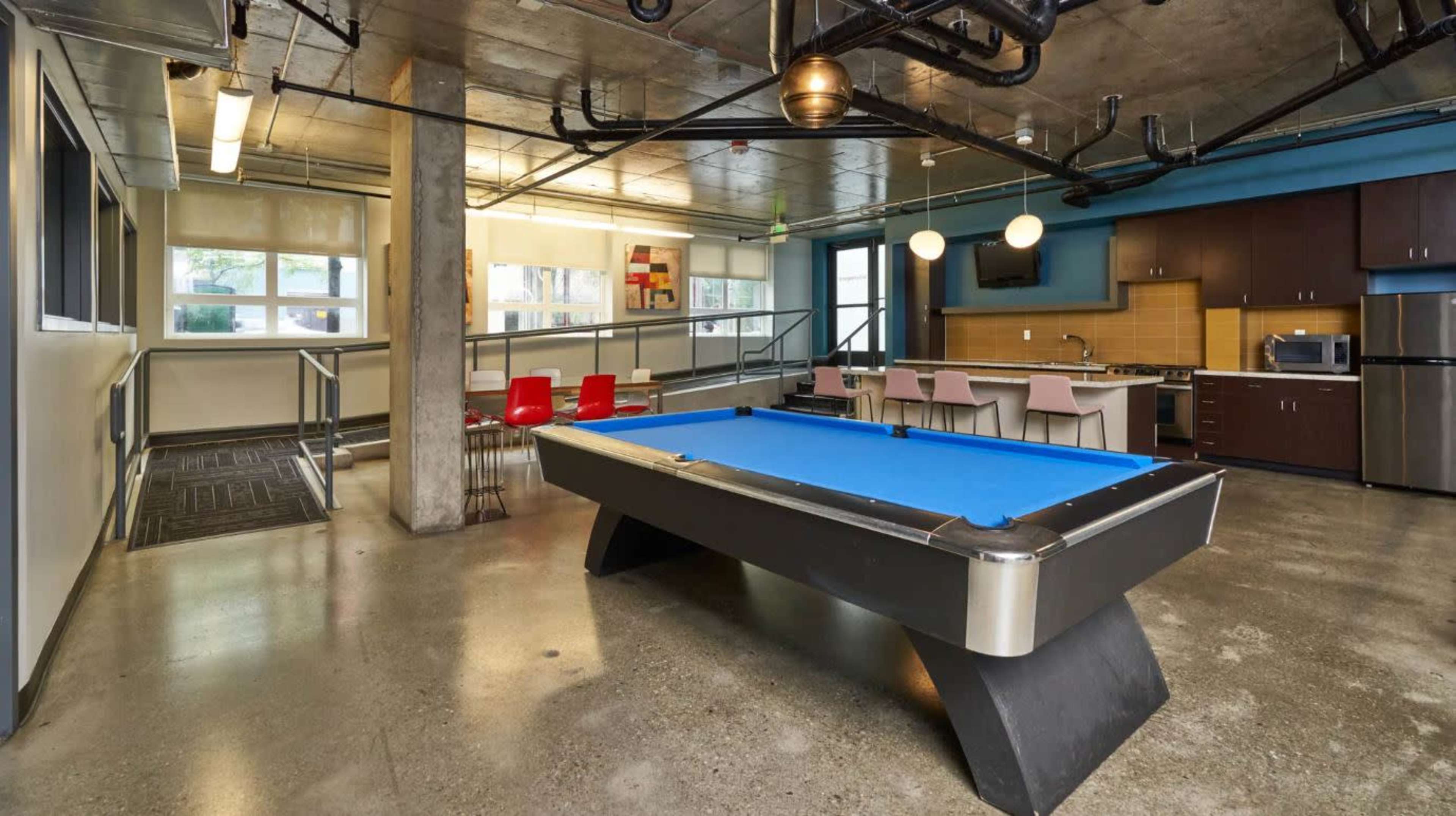 The image shows a modern game room with a blue pool table, a kitchenette, and seating areas.