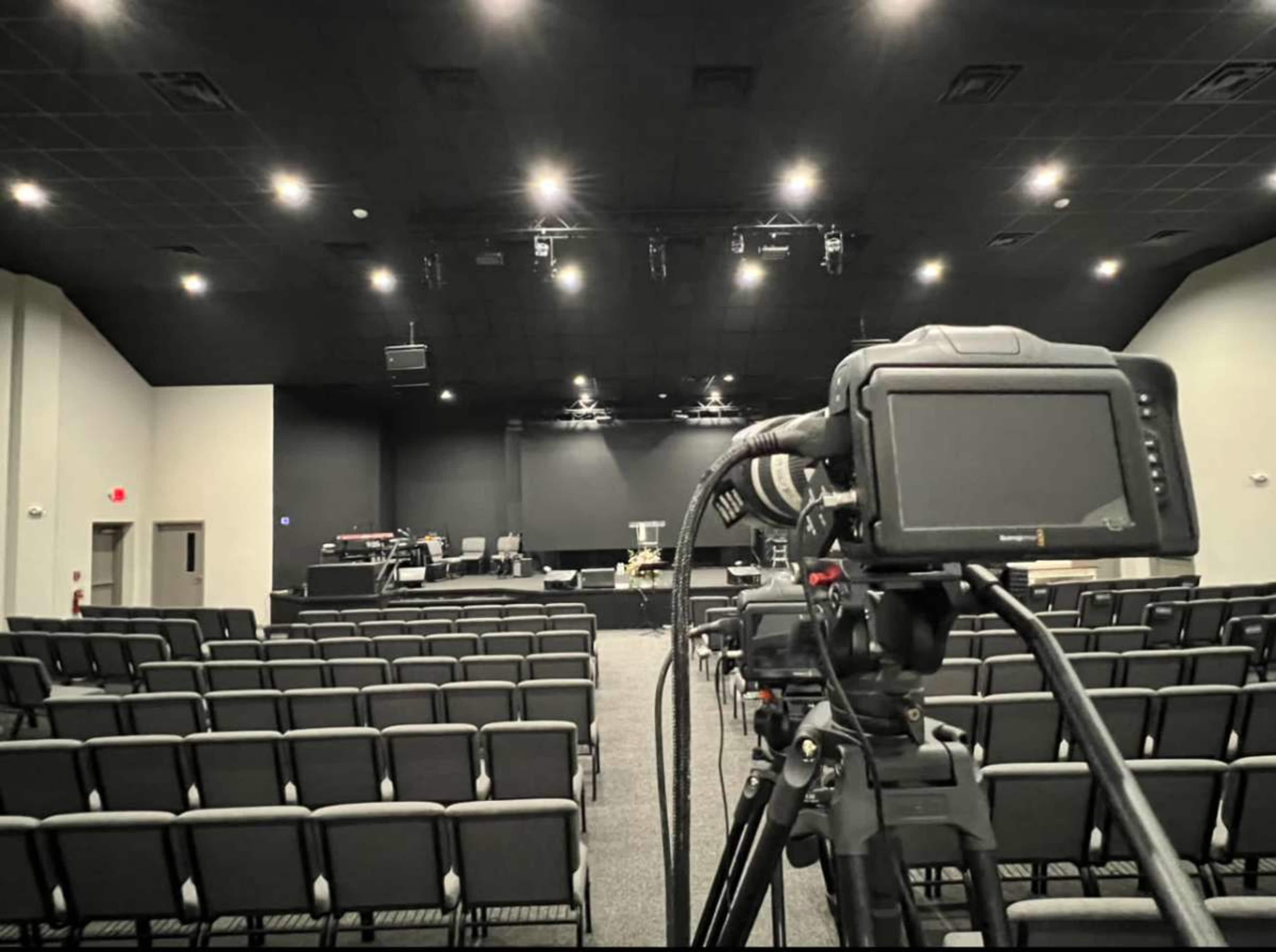 A camera is positioned in front of empty rows of seating facing a stage in a darkened auditorium.