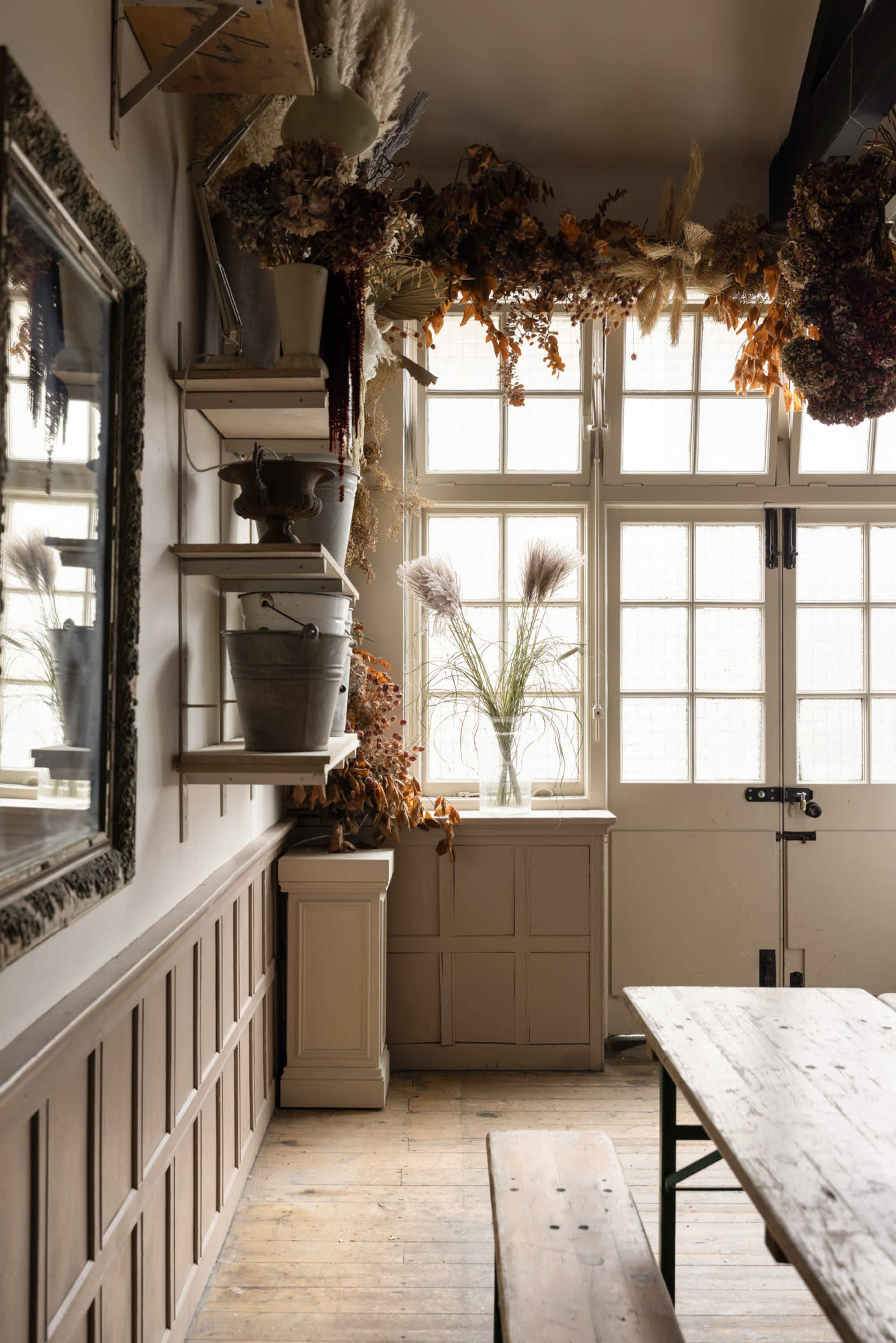 The image shows a cozy interior with wooden flooring, a long, simple table, and dried flowers arranged on shelves and window sills.