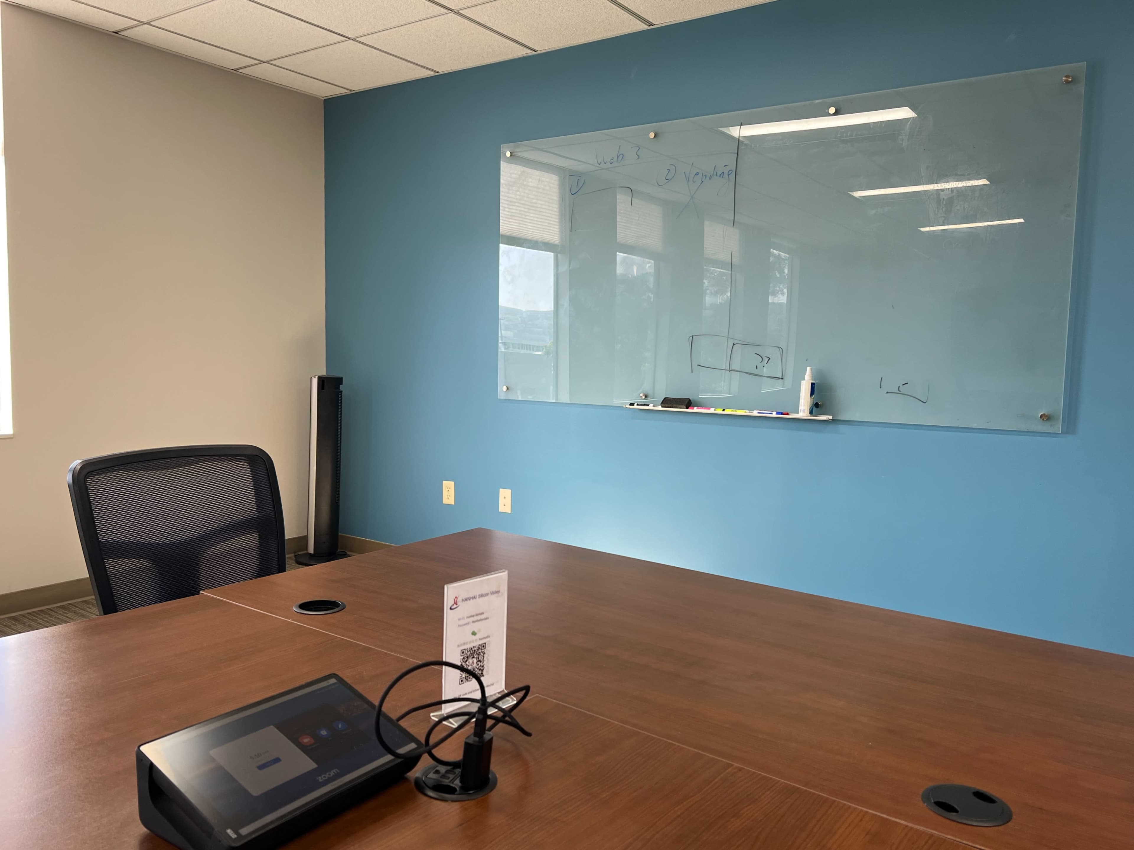 A conference room with a wooden table, a black chair, a digital device on the table, and a glass whiteboard mounted on a blue wall.