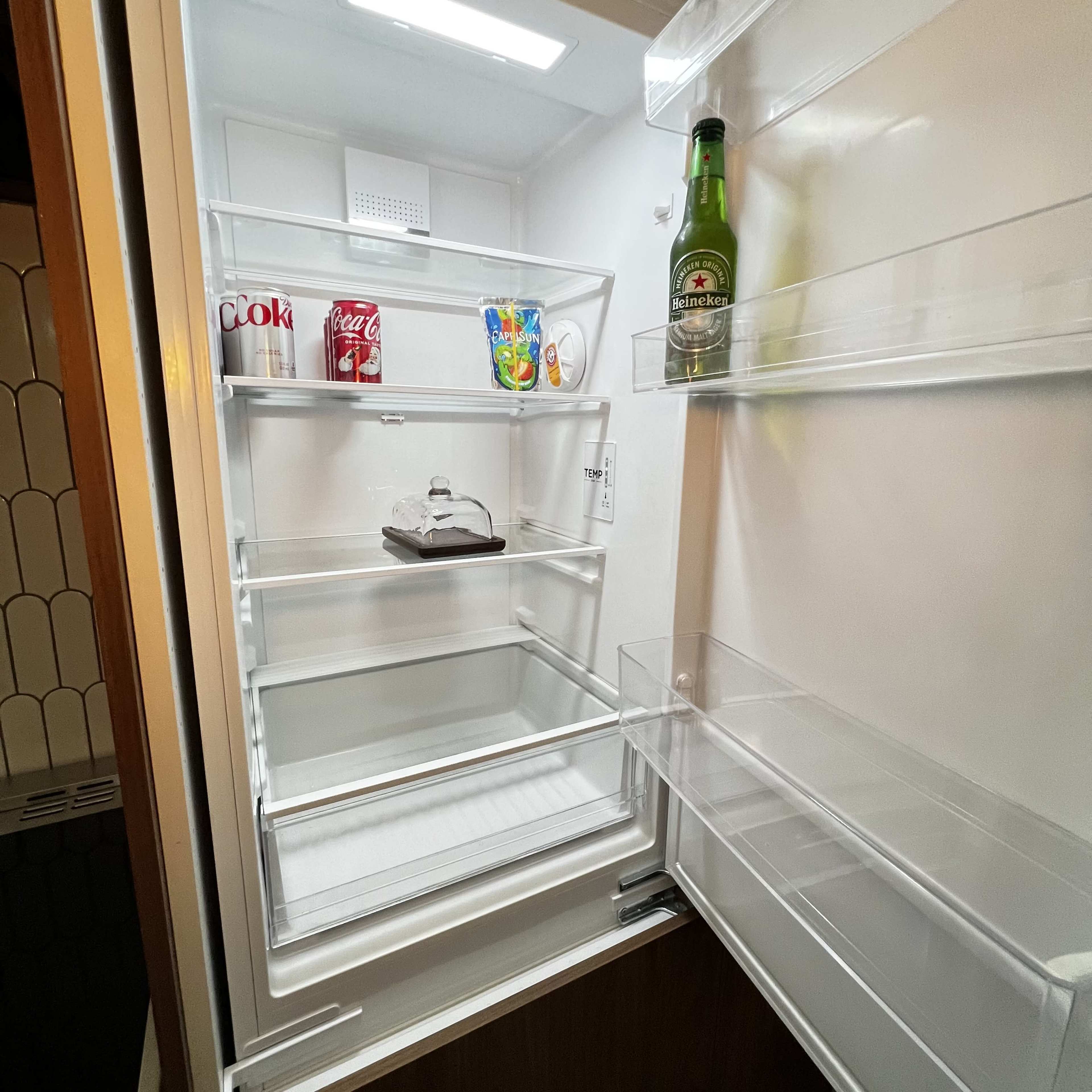The interior of a refrigerator shows a few cans of soda, a bottle of beer, a small dish, and some partial items on empty shelves.
