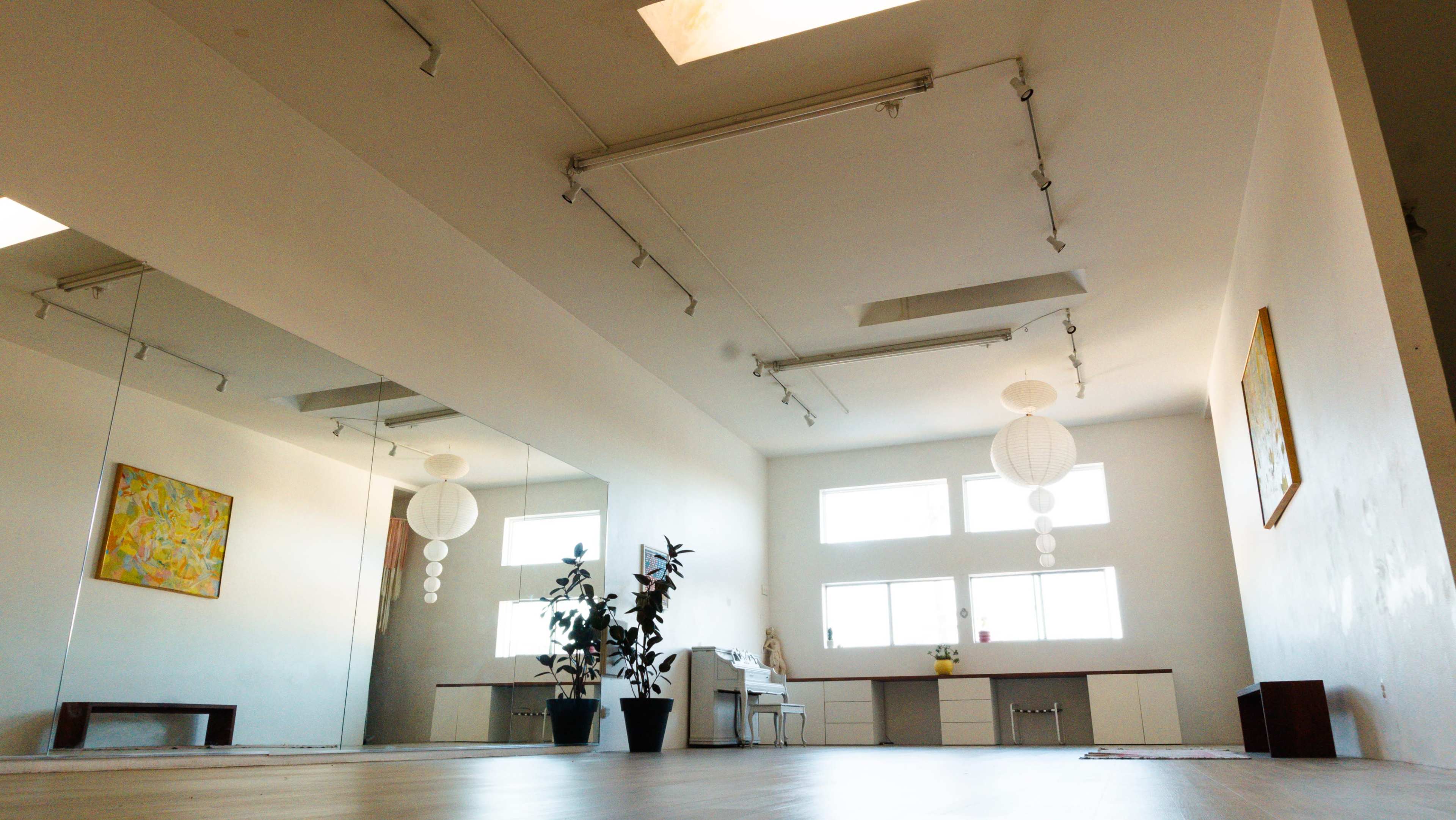 The image shows a spacious room with a wooden floor, large mirrors on one wall, and minimal decor, including a plant and a few hanging light fixtures.