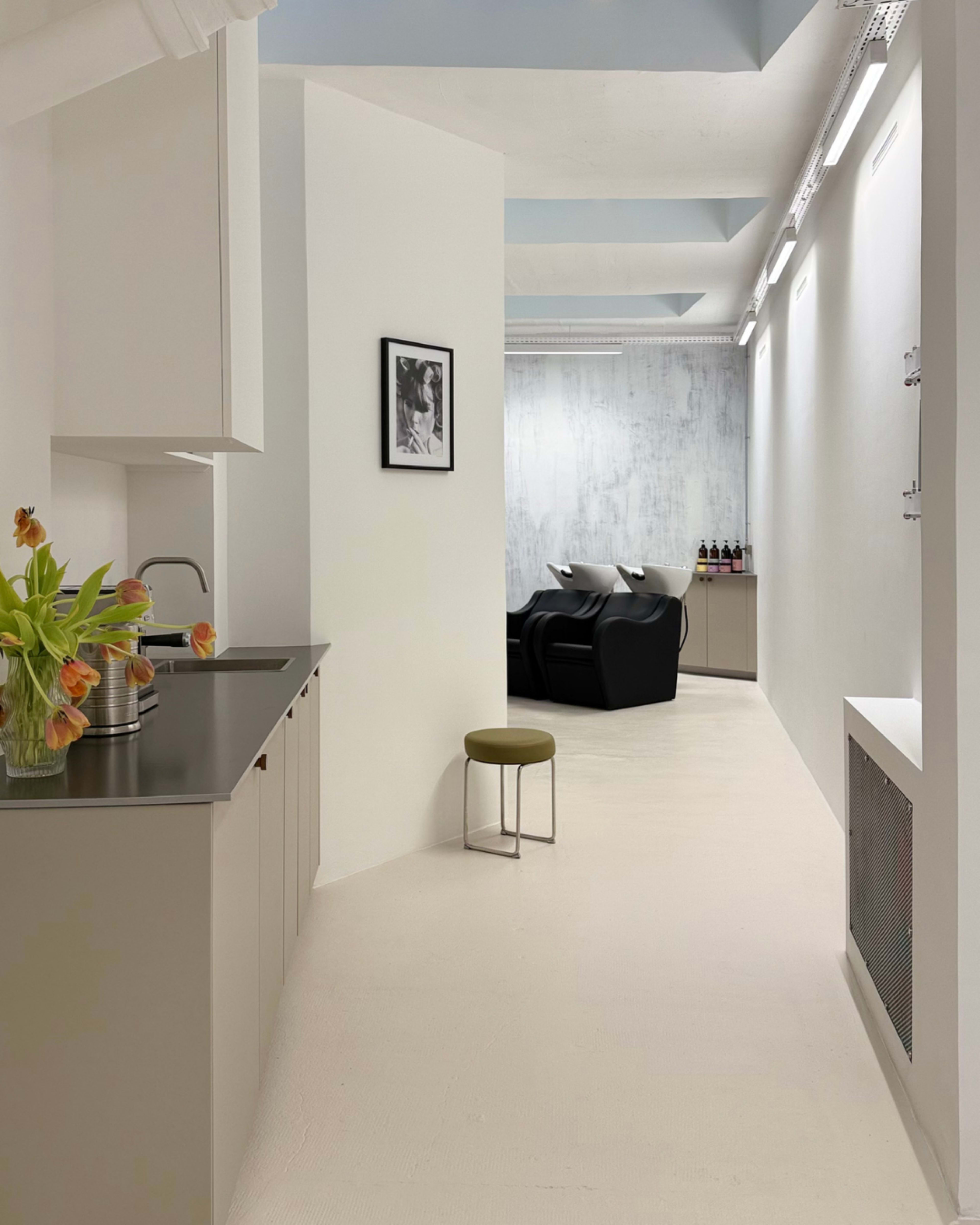 The image shows a modern interior space featuring a kitchen area on the left, with flowers on the counter, and a seating area with two black chairs in the distance.