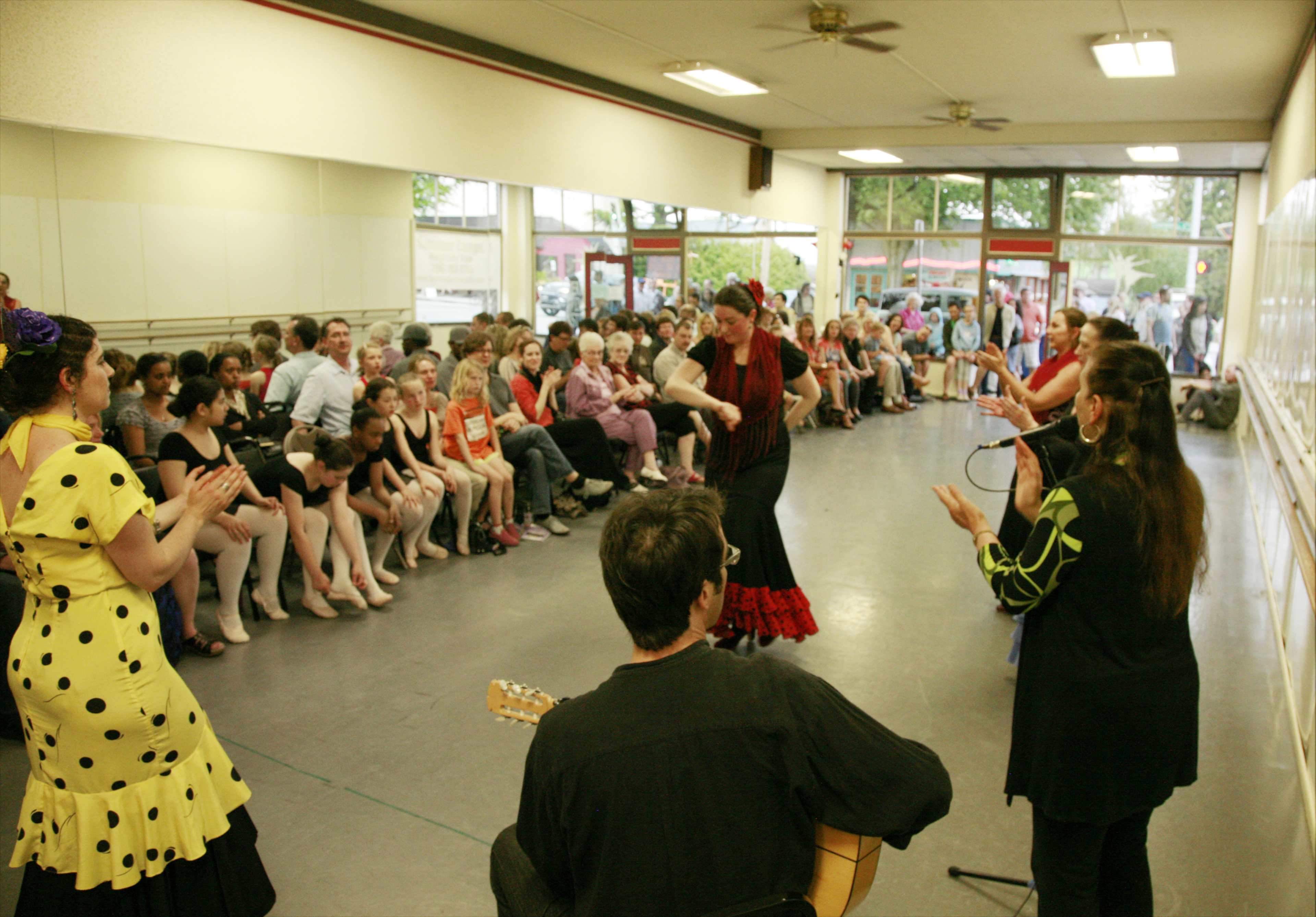 A group of dancers perform in front of an audience seated in a dance studio.