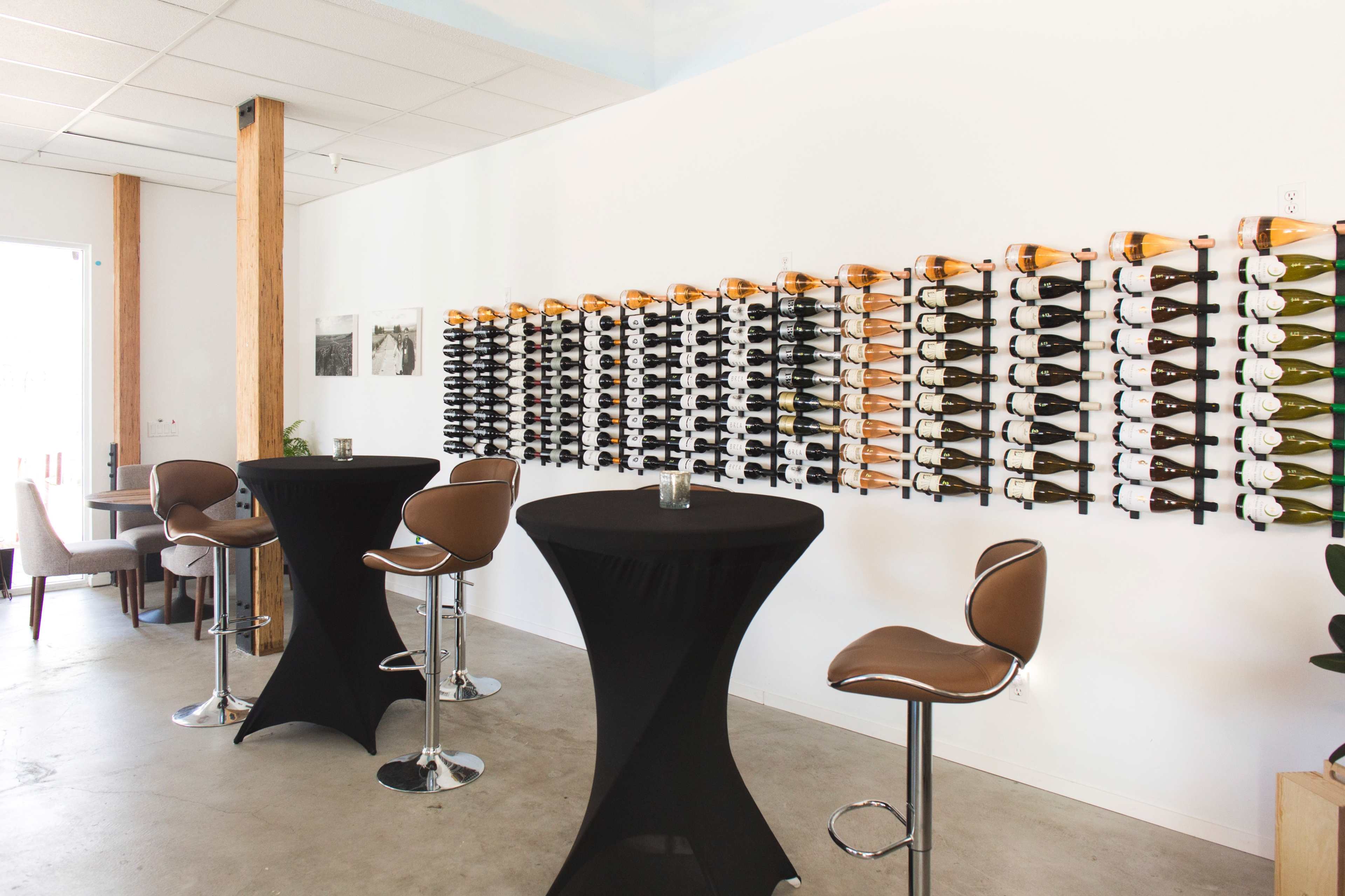 A modern interior of a wine bar with a wall-mounted wine rack displaying various bottles and several high-top tables with black tablecloths.