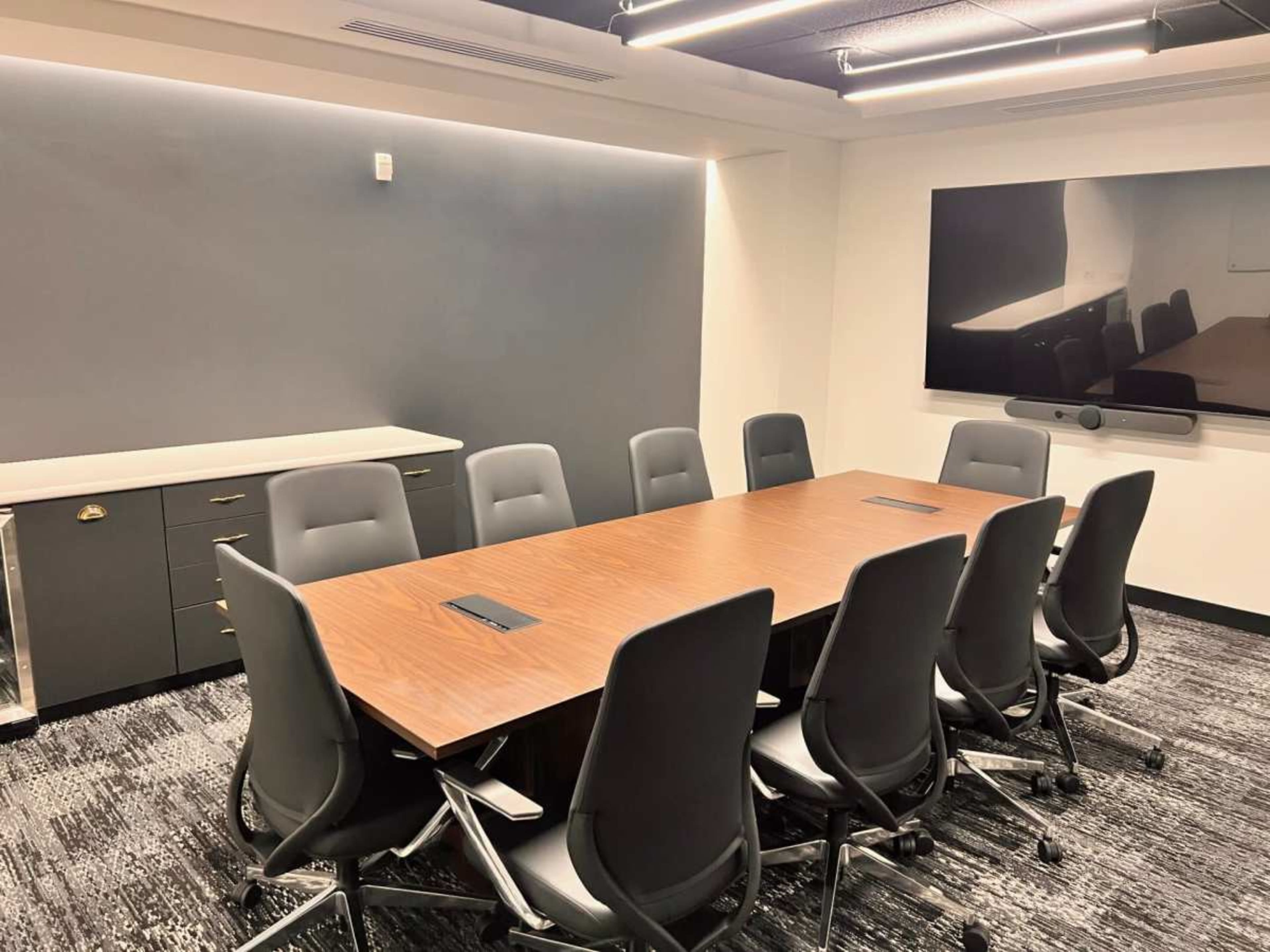 A conference room features a long wooden table surrounded by ten black ergonomic chairs, with a large screen mounted on the wall at one end.