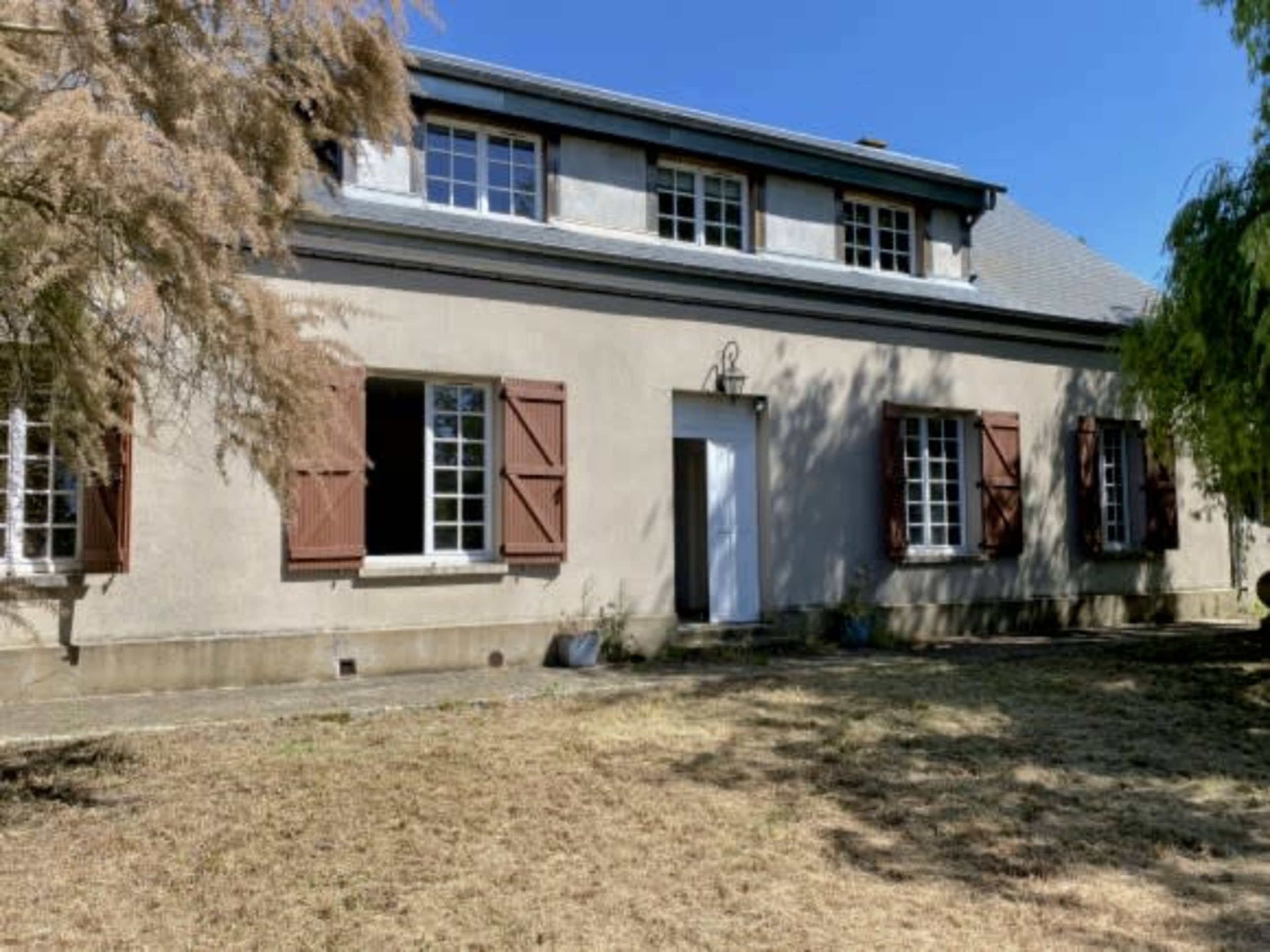 A two-story house with shuttered windows and a white front door is surrounded by a dry grassy area.