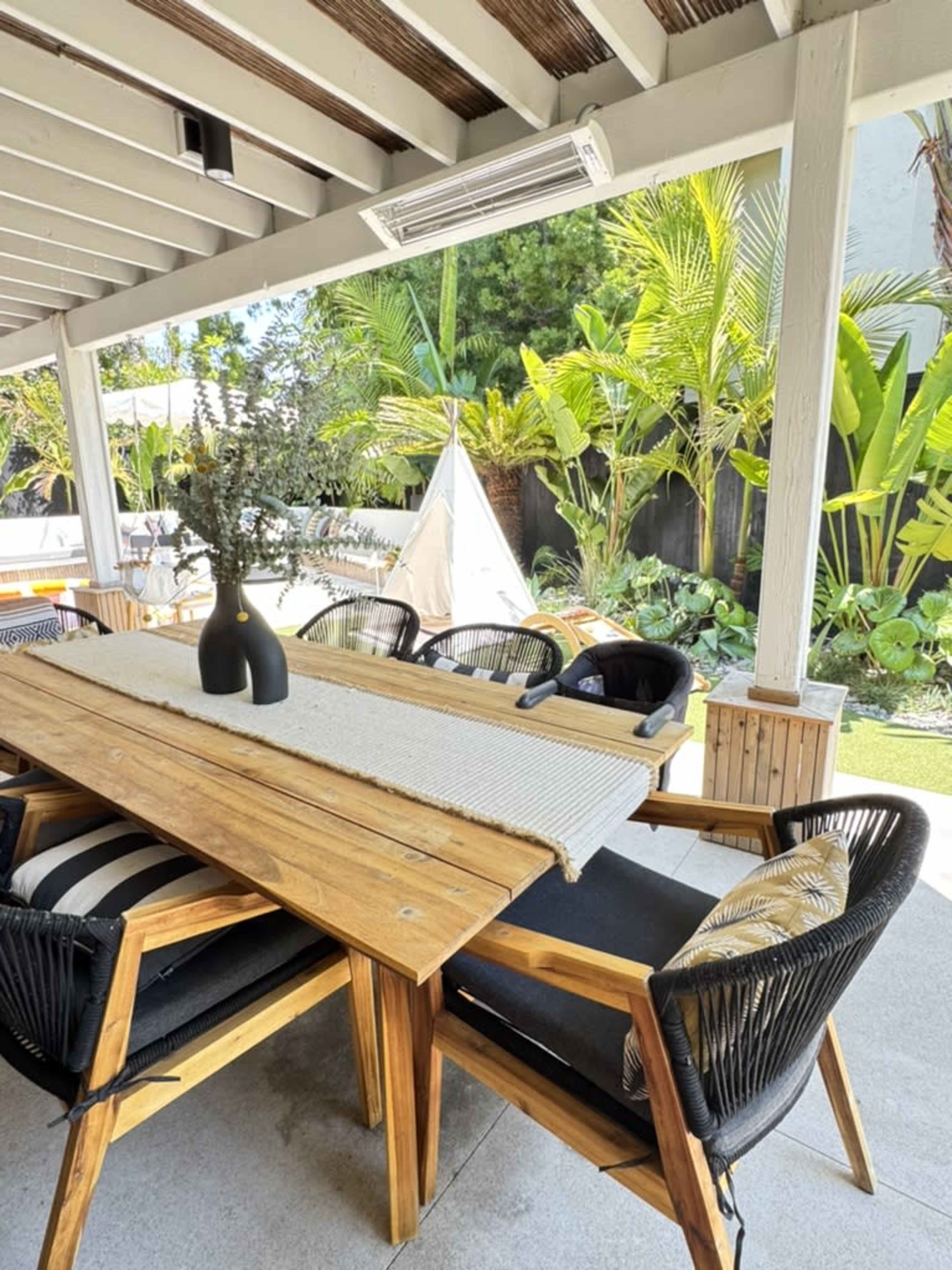 A wooden dining table is set under a covered patio, surrounded by black chairs and tropical plants in the background.