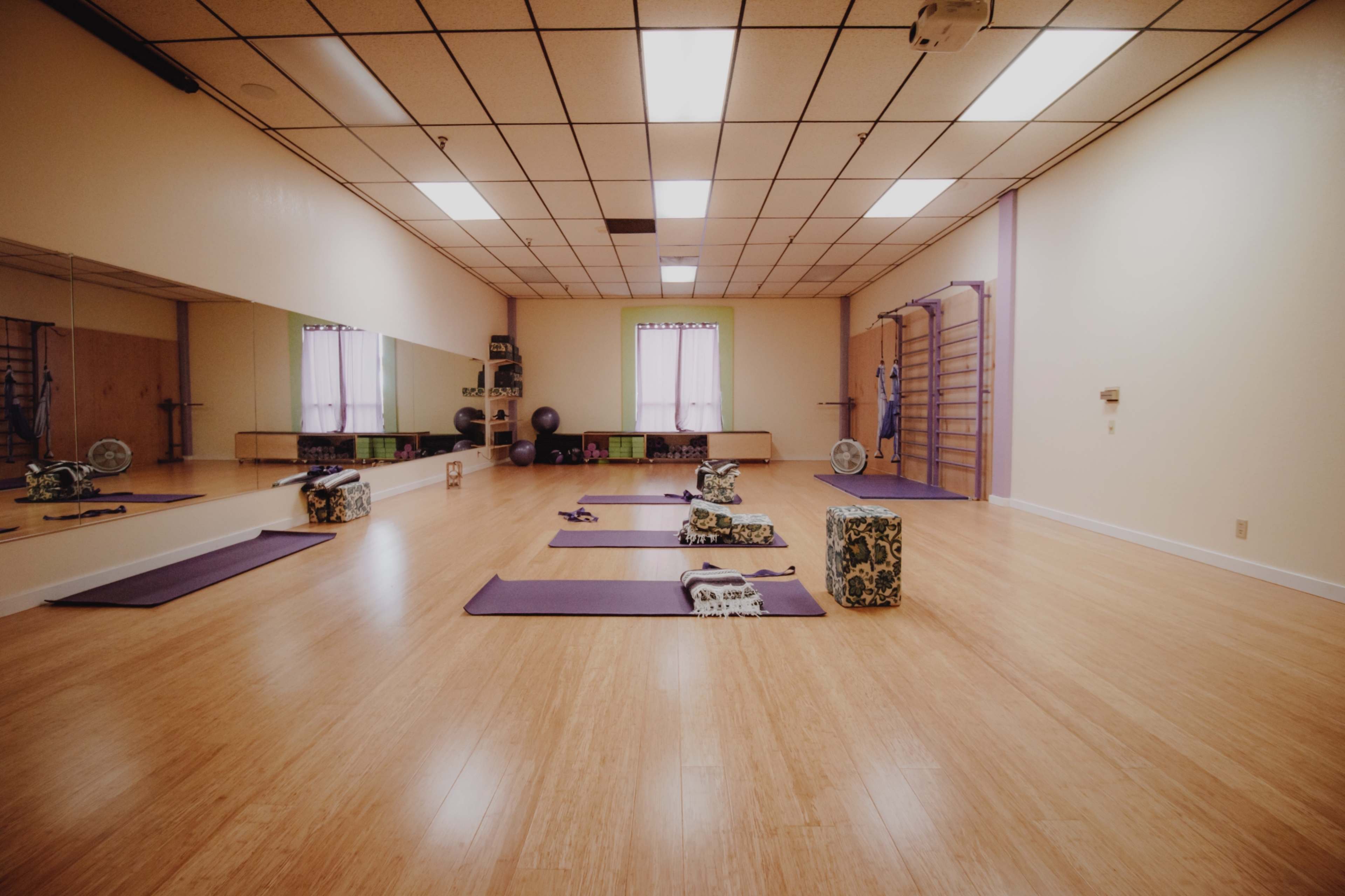 The image shows a spacious yoga studio with purple mats arranged on a bamboo floor, a large mirror, and various exercise equipment along the walls.