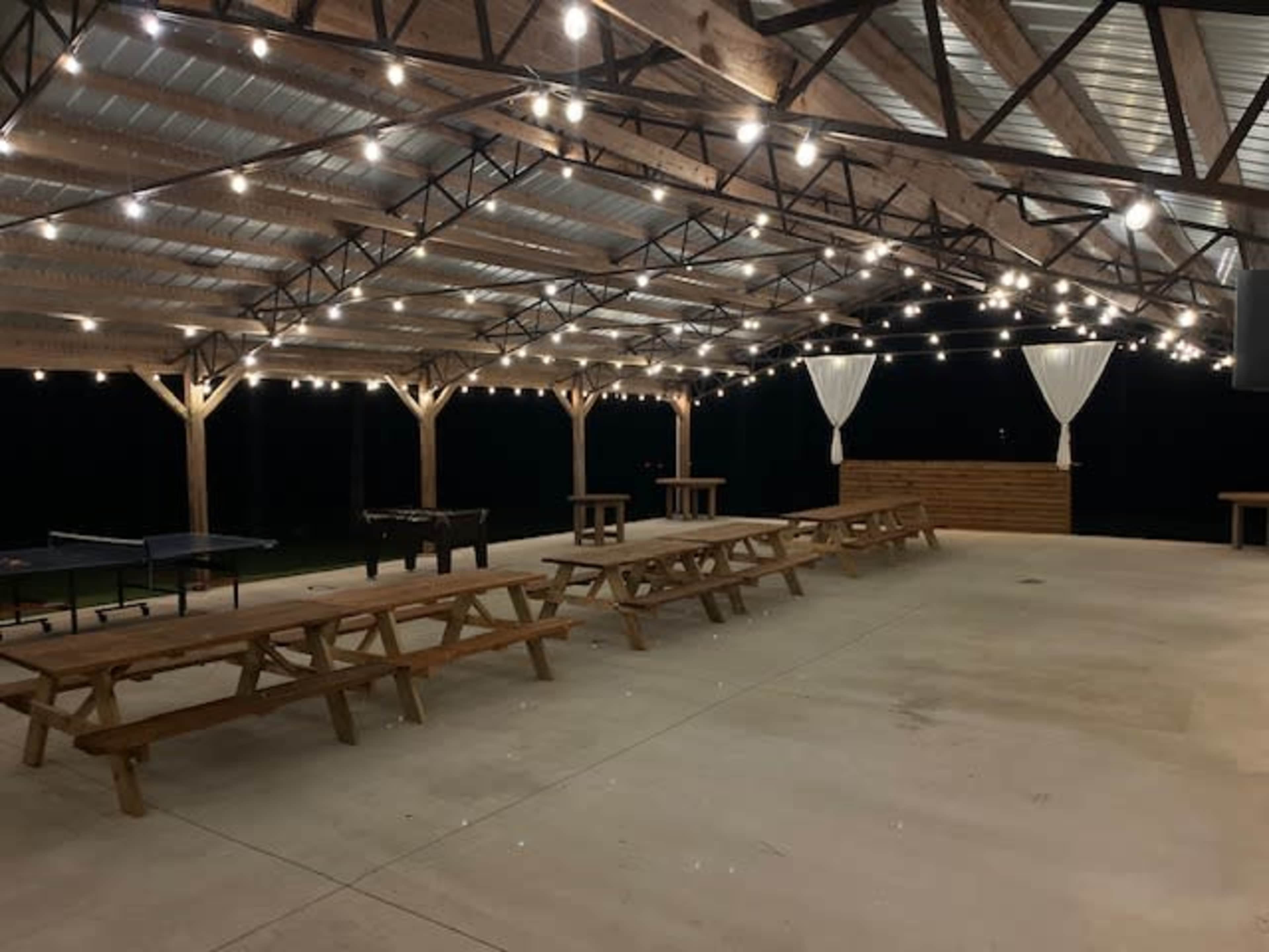 An outdoor pavilion with string lights, wooden picnic tables, and a bar area, set against a dark background.